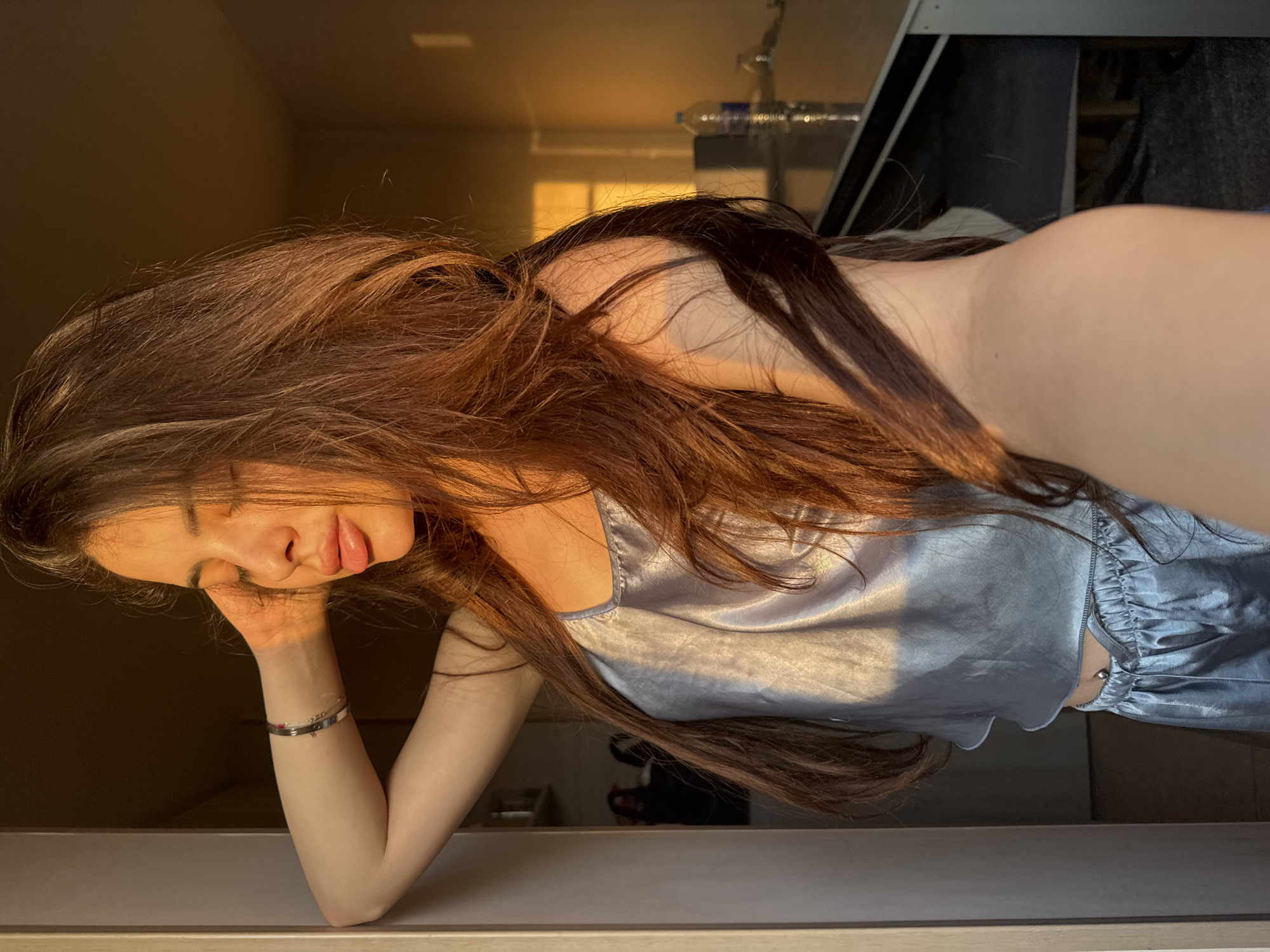 A young adult female is captured in a residential interior in Plovdiv, Bulgaria, illuminated by warm, golden hour sunlight. She is seen from the chest up, leaning with her right elbow resting on a light-colored vertical door frame or wall. Her right hand is positioned gently near her temple, and her eyes are closed with her lips slightly parted, suggesting a moment of repose. She wears a two-piece pajama set made of light blue or silver satin, comprising a camisole and matching shorts, with a silver belly button piercing visible at her midriff. A silver bracelet is worn on her right wrist. Her long, dark brown hair, with auburn tones accentuated by the light, cascades over her shoulders and chest. The image is a selfie, taken from a low angle. In the background to her right, a reflective dark surface, possibly a table or counter, holds a plastic water bottle and a glass. A distinct rectangular patch of golden light, indicating a window off-frame, is cast onto the distant wall, creating prominent shadows within the warmly lit room.