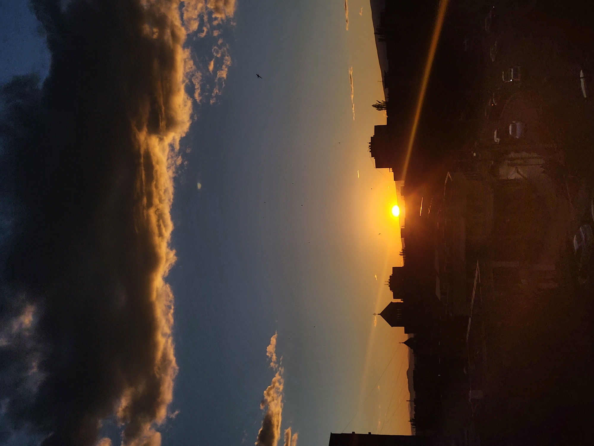 The image captures a sunset over Yerevan, Armenia. Silhouetted buildings, including a church with a prominent cross, are visible against the bright setting sun. Large, dark clouds with golden edges flank the left side of the frame. A few birds are visible in the sky. The right side of the image is darker, showing more buildings and possibly parked vehicles. The overall scene is one of urban sunset.