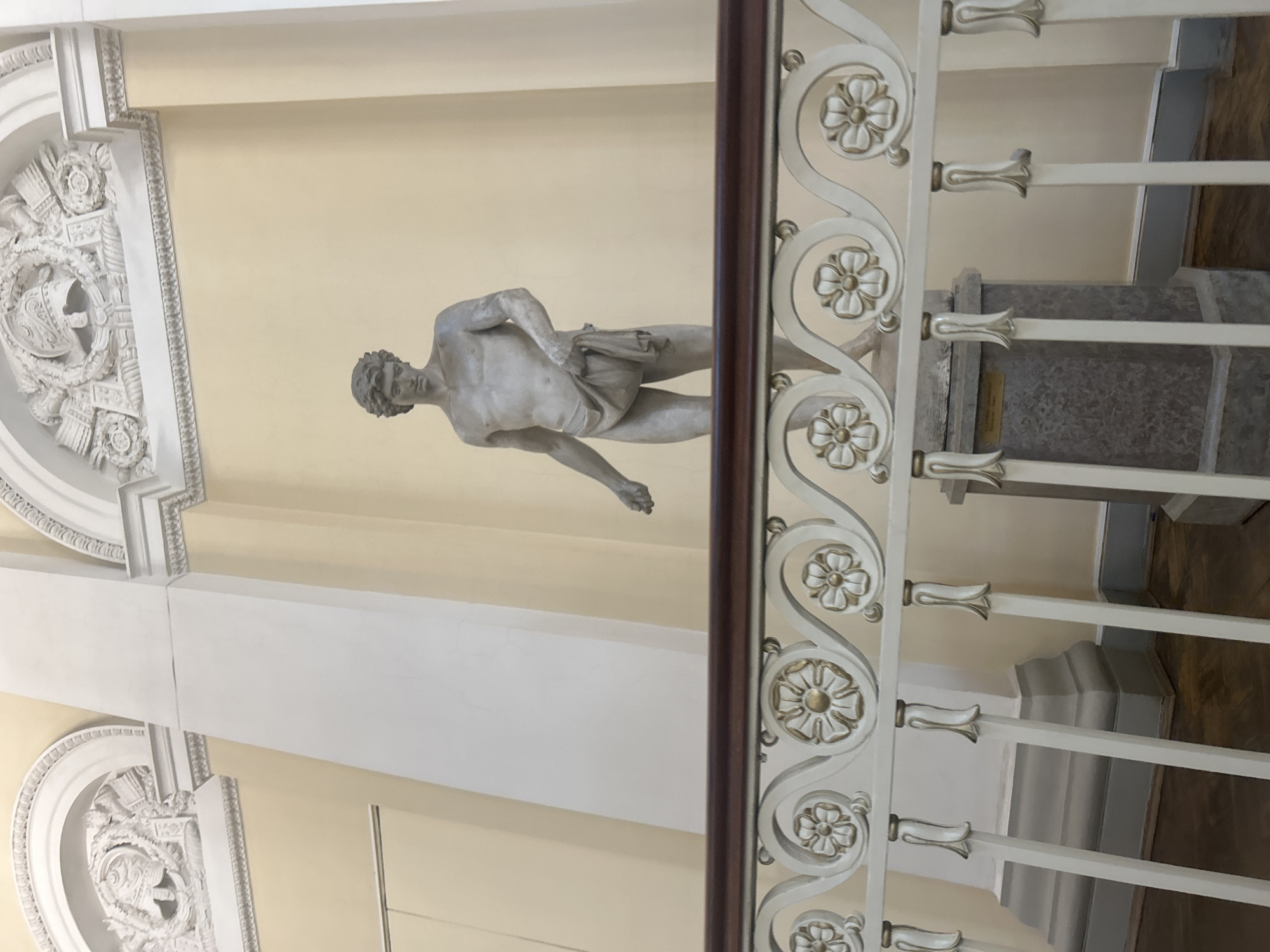 A full-body, eye-level, outdoor shot shows a pale, stone statue of a young man wearing a draped cloth. The statue stands on a grey, marbled pedestal in the center of the frame, against a pale yellow wall. Decorative white and gold railings with floral motifs are in the foreground, obscuring the lower portion of the scene. Architectural details like white columns and arched recesses with decorative reliefs are visible in the background. The lighting is diffused, suggesting an overcast day or an indoor setting with indirect light. The overall scene is one of classical architecture and art.