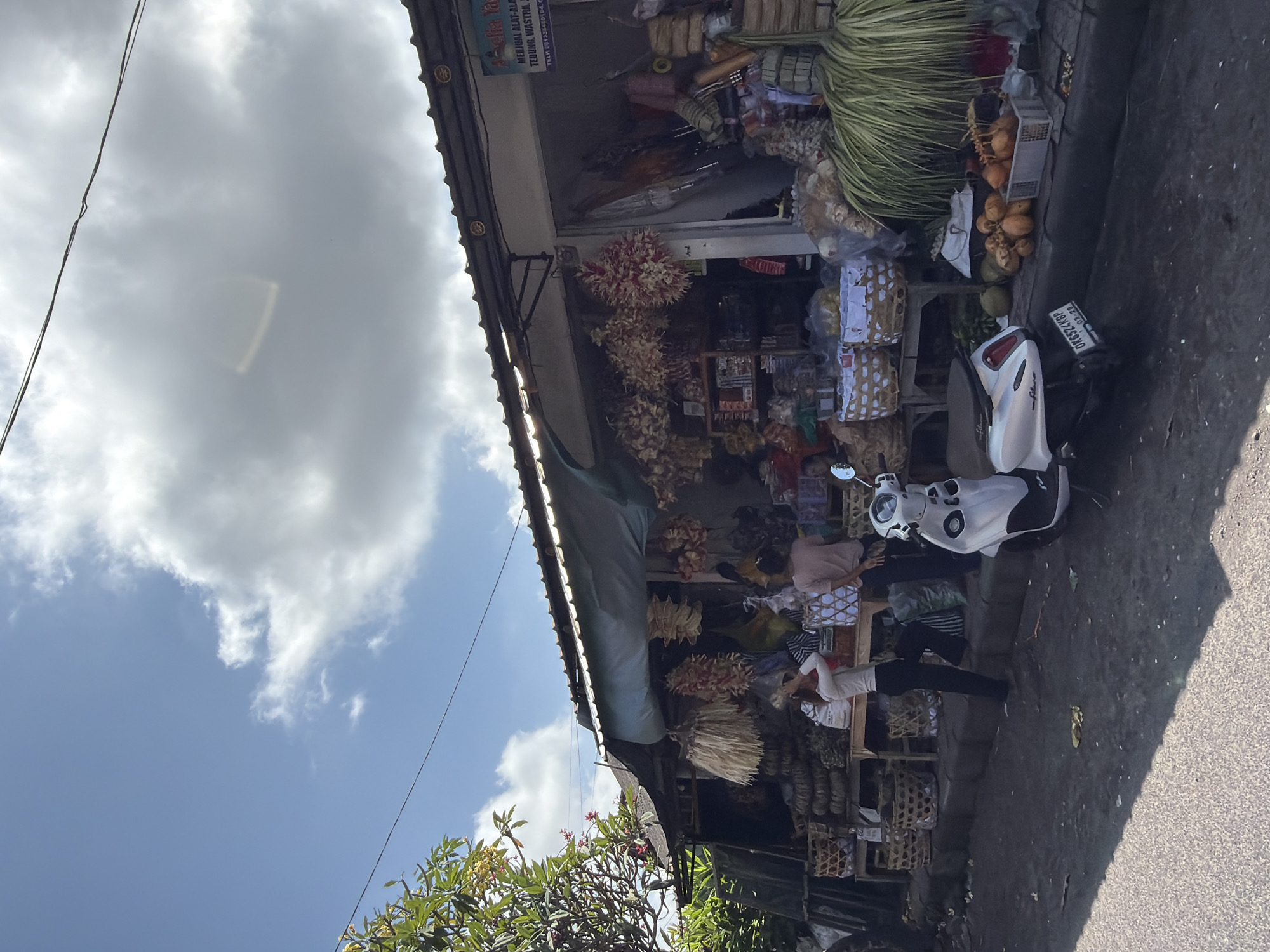 The image depicts a street scene in Blahbatuh, Indonesia, featuring a market stall selling various items, likely dried goods and crafts. In the foreground, a white scooter is parked on the side of the road. A woman in a white long-sleeved shirt and black pants stands near the market, adjusting her hair. Another person, wearing a pink t-shirt and dark pants, stands beside her. The market stall itself is covered by a blue awning and displays an assortment of goods, including straw-like decorations hanging from the roof, baskets filled with items, and what appear to be coconuts and other produce arranged on the ground. The street in front of the market is paved, and the area around it is somewhat dusty. The sky is blue with scattered white clouds.