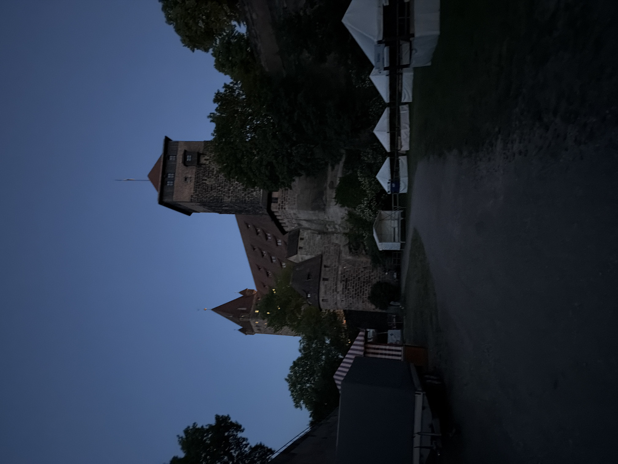 The image shows the Nuremberg Castle in Nuremberg, Germany at dusk. The castle's towers and red tile roofs are visible against a darkening blue sky. Trees and vegetation surround the castle walls. Several white tents are lined up along a pathway adjacent to the castle. A dark, paved path stretches into the distance. In the foreground, the corner of a dark, modern structure is visible, along with a red and white striped covering.