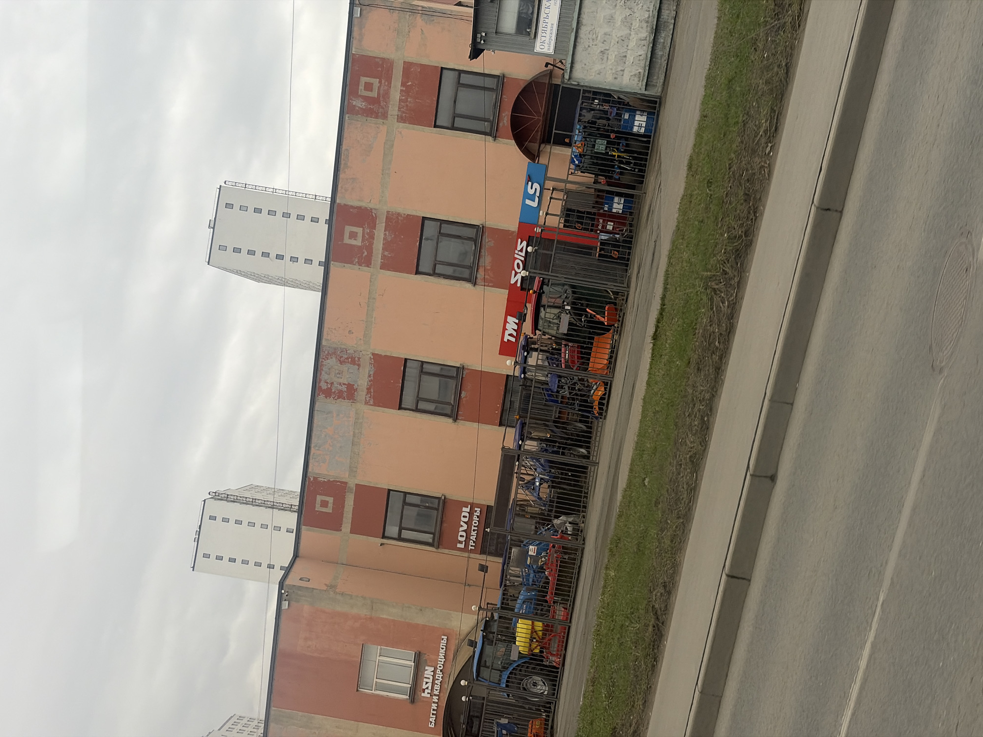 The image shows a building in Saint Petersburg, Russia that appears to be a dealership for tractors and ATVs. The building is peach-colored with brown brick accents and multiple windows. A sign on the building displays brands such as LS, Solis, TM, LOVOL, and HSUN. The street is visible next to the building, along with a grassy area and a sidewalk. In the background, several high-rise apartment buildings can be seen. The sky is overcast.