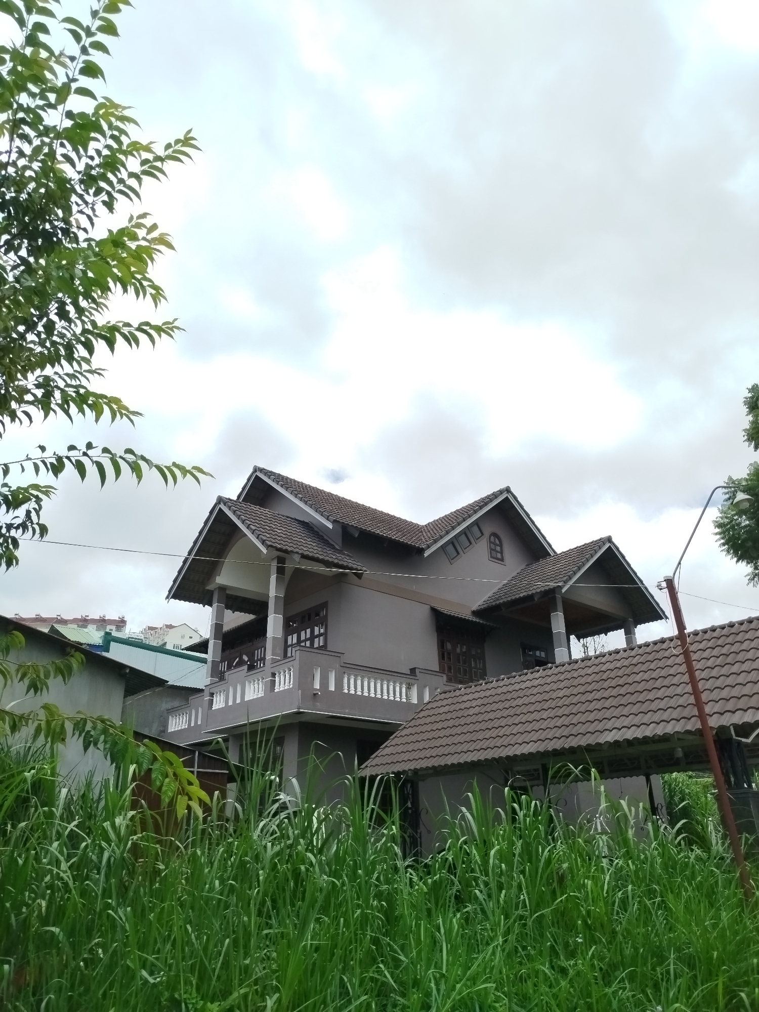 The image captures a two-story house in the city of Da Lat, Vietnam, characterized by its gray stucco exterior and dark tile roofing. The house features multiple gables, balconies with white railings, and large windows. Tall, lush green grass dominates the foreground, partially obscuring the lower level of the house and a covered patio area. A single lamppost is visible on the right side of the frame. The sky is overcast with heavy clouds. The surrounding environment appears to be a residential area with other buildings visible in the background, including structures with red tile roofs. The scene is static and devoid of human activity.