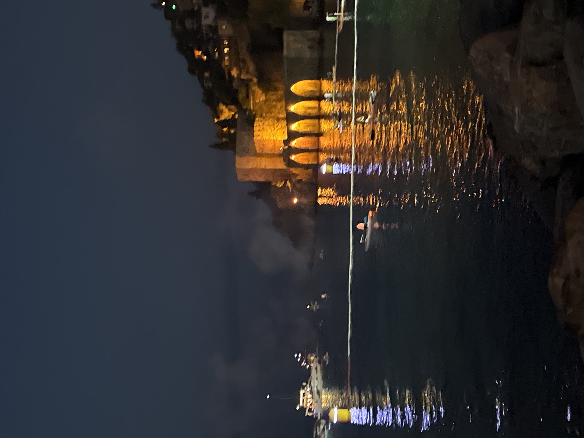 The image captures a nighttime scene of a coastal city, likely Alanya, Turkey, given the context. The foreground is dominated by dark water reflecting artificial lights, creating streaks of yellow, orange, and purple. On the right side of the frame, large rocks are visible, adding to the natural landscape.
In the mid-ground, a boat with a person on it is visible in the water. A thin white rope extends from the boat towards the right side of the frame. Further back, a fortified wall with several arched openings, illuminated by warm yellow lights, stands imposingly. These arches are reflected in the water below. Above the wall, the silhouettes of buildings with scattered lights suggest a town or city built on a hillside. The overall atmosphere is serene and illuminated, characteristic of a city at night. The time of day is clearly night, with a dark sky and artificial lighting creating the scene. There is no visible text in the image.