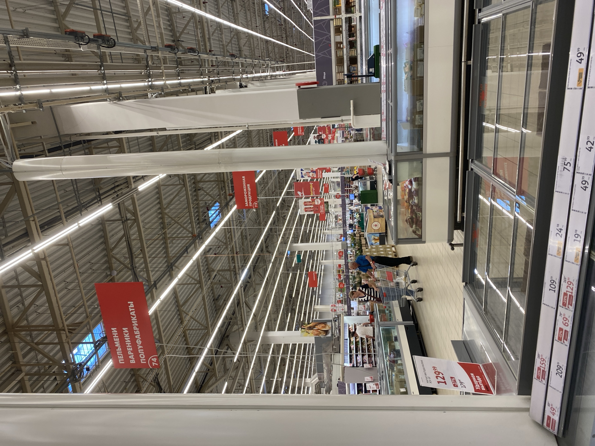 This image shows the interior of a supermarket in Kuzmolovskiy, Russia. The camera is positioned at eye level, looking down an aisle filled with refrigerated display cases. In the foreground, a row of empty, glass-lidded freezer units is visible. Further back, several people are shopping, including a couple pushing a shopping cart and another person looking at products in a refrigerated case. Overhead, fluorescent lights illuminate the expansive ceiling, which is crisscrossed with industrial beams and pipes. Red signs with Russian text hang from the ceiling, indicating different product sections, such as "PELMENI VARENIKI POLUFABRIKATI" (dumplings, vareniki, semi-finished products) and "ZAMOROZHENNAYA PRODUKTSIA" (frozen products). The overall environment appears clean and well-lit, typical of a large grocery store.