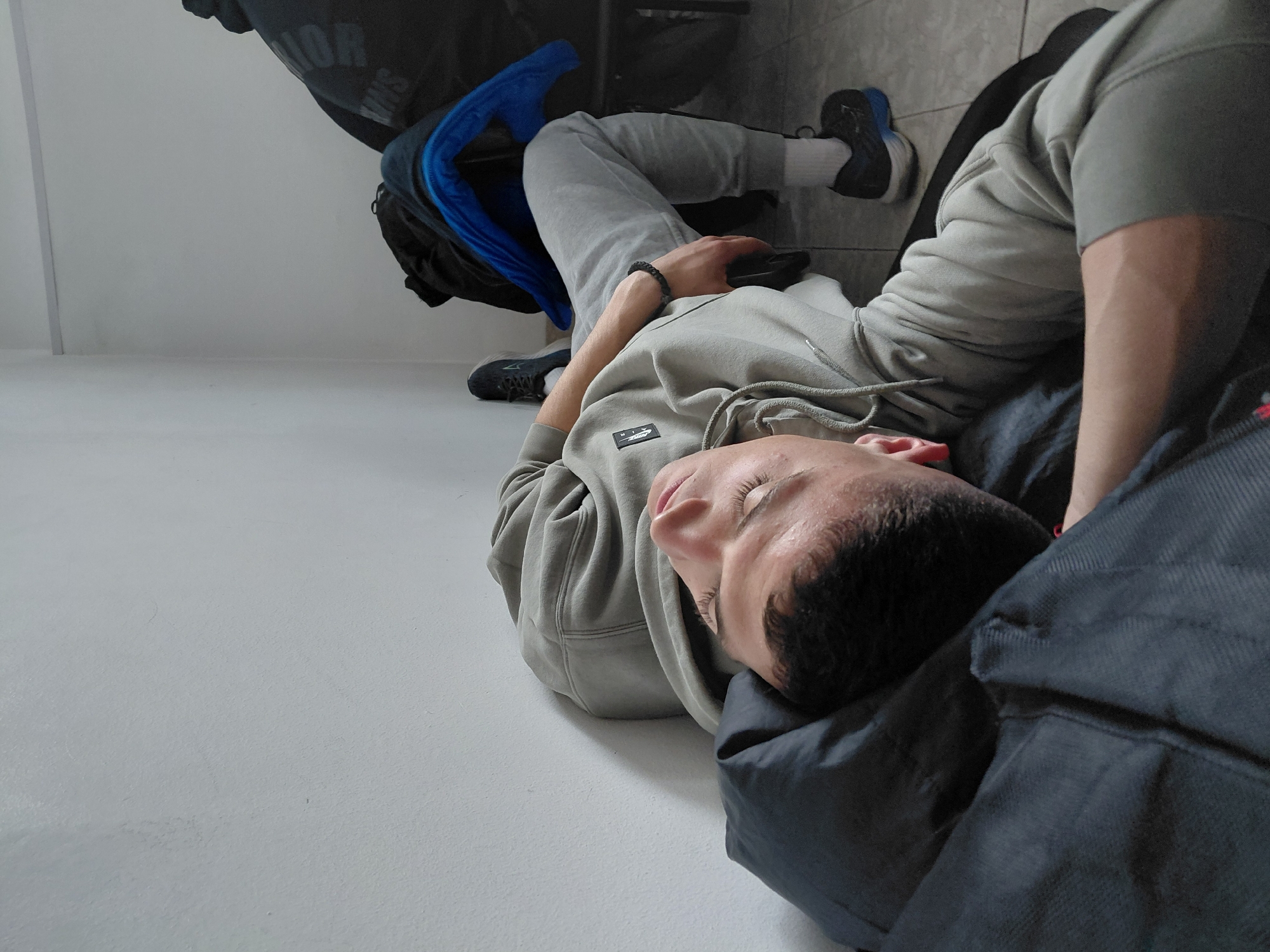 A young man lies on his side on a light gray floor. He is wearing a gray hooded sweatshirt and gray sweatpants, with white socks and black sneakers with blue accents on his feet. His head is resting on a dark blue or black bag. Another bag, also black, is behind him. His eyes are closed, and he appears to be sleeping. He has a bracelet on his left wrist and is holding a black object, possibly a phone, in his hand. The scene is indoors, with a light gray or white wall in the background. There is no explicit evidence to definitively confirm the location in Sofia, Bulgaria based solely on the visual information provided in the image. The location is unknown.