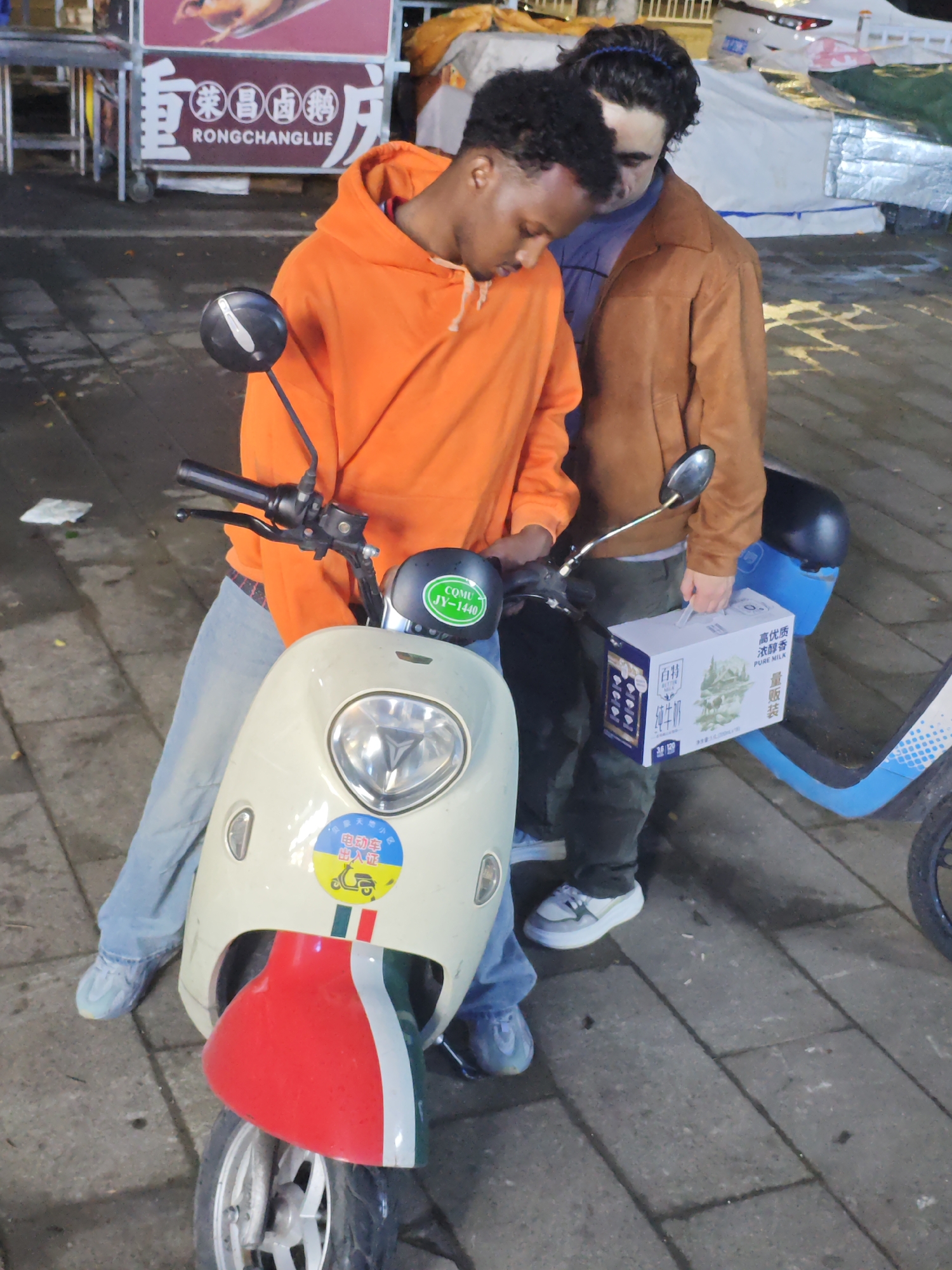 A medium shot captures two men standing next to parked scooters on a paved surface. The man on the left is dark-skinned, wearing an orange hoodie, light blue jeans, and light blue sneakers. He appears to be looking down at the scooter in front of him. The scooter has a white body, a red and green fender, and a sticker with Chinese characters and a small scooter graphic on the front. To his right, the second man, who appears to be light-skinned, is wearing a brown jacket, green pants, and white and gray sneakers. He is holding a white box with Chinese characters and a picture of a landscape.
To the right of the first scooter is a second scooter, which is predominantly blue. In the background, a food stall or cart is visible with Chinese characters and the English words "RONGCHANGLUE" written on it.
The scene seems to be a street in the city of Chongqing High-tech Zone, China. Several stickers with Chinese characters and what appears to be information relevant to an electric scooter are visible on the scooter.