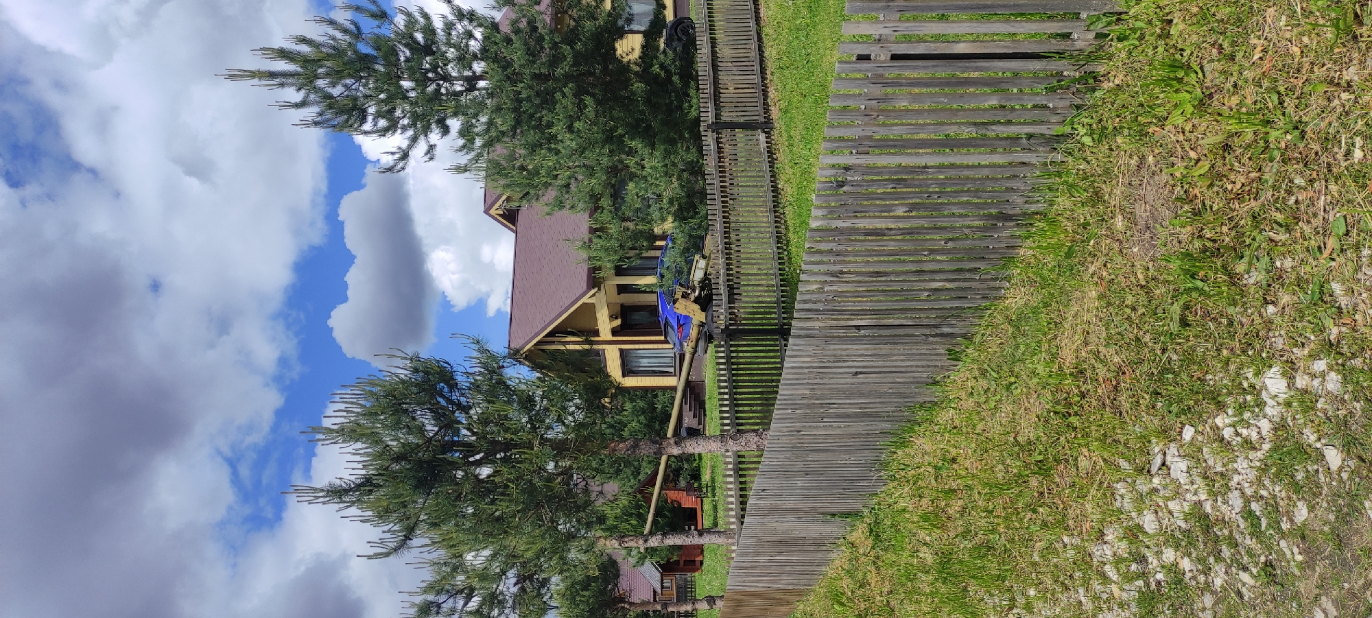 A sideways outdoor shot shows a yellow house with a brown roof behind a wooden fence, with a partially cloudy sky in the background. The house is two stories and the fence runs along the border of the property. Green grass and trees are also visible. The location is Ivanovskoe, Russia.