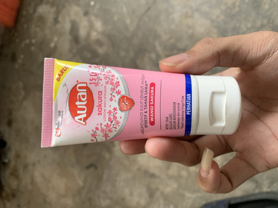 A person's hand holds a tube of Autan Sakura mosquito repellent lotion, oriented horizontally against a blurred, light grey concrete or tiled background. The hand has a light to medium brown skin tone, with noticeably long fingernails on the index finger.
The pink and white tube is clearly visible, featuring the brand name "Autan" in red, along with "sakura" and "LOSION ANTI NYAMUK" (Mosquito Repellent Lotion). A yellow banner on the top left corner reads "BARU" (New). The packaging is decorated with delicate pink sakura (cherry blossom) flowers and a graphic of a red fruit, possibly a strawberry or lychee.
Visible text on the tube includes:
- "MELINDUNGI KULIT LEMBUT ANDA*" (Protects your soft skin*)
- "EFEKTIF & TAHAN LAMA**" (Effective & Long-lasting**)
- "WANGI SAKURA" (Sakura Scent)
- "NETTO: 50ml" (Net: 50ml)
- "BACALAH LABEL SEBELUM MENGGUNAKAN" (Read label before use)
- "*perlindungan dari nyamuk" (*protection from mosquitoes)
- "**Efektif melindungi hingga 8 jam" (**Effectively protects for up to 8 hours)
- "PERHATIAN" (Caution/Attention)
- A small "S.C. Johnson & Son, a family company" logo is also visible.
The product has a white flip-top cap on the right. The lighting suggests daytime. The scene provides no specific emotional cues. The setting appears to be an indoor or outdoor area with a simple, utilitarian floor surface, typical of Metro, Indonesia.