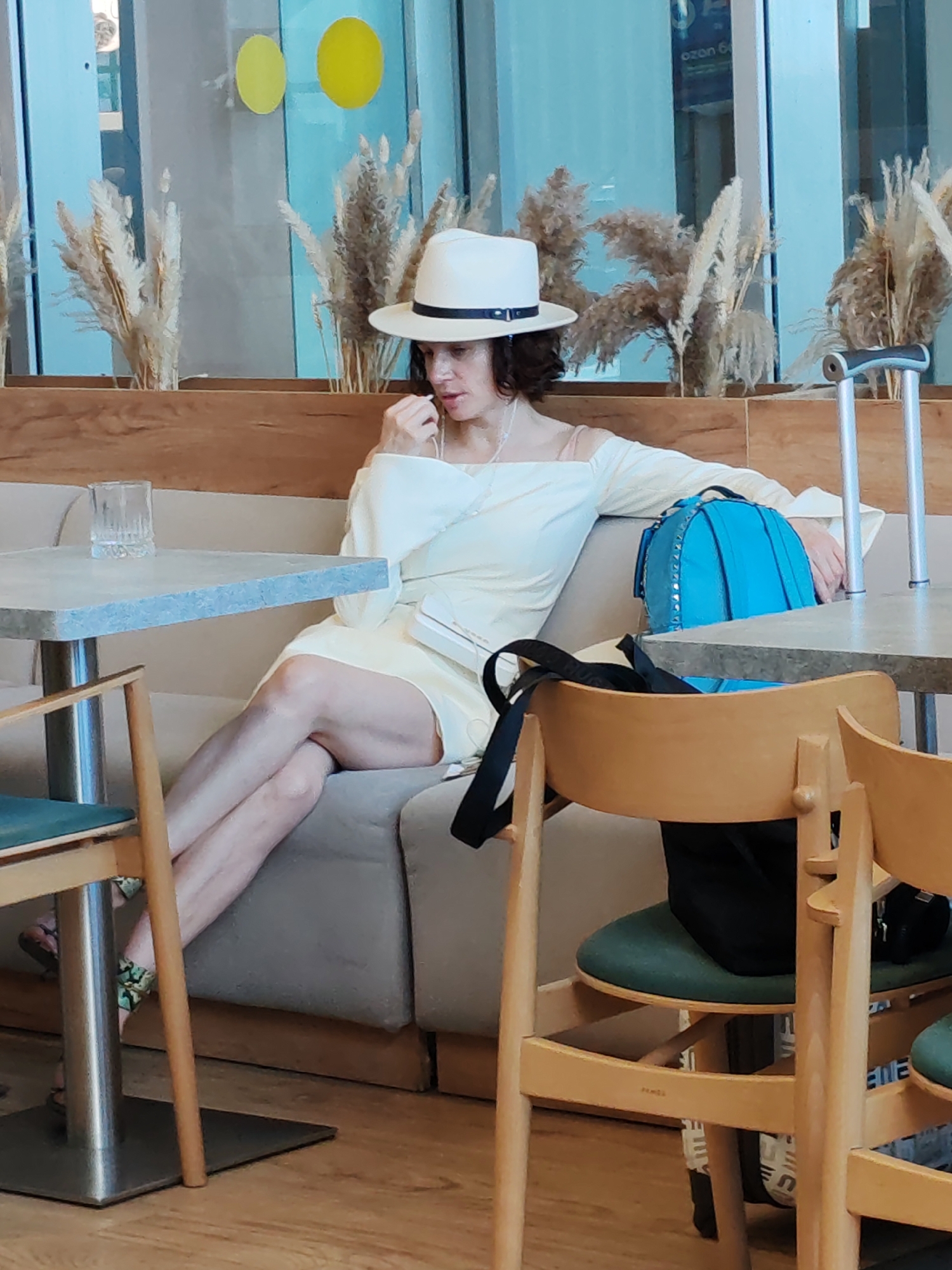 In a brightly lit indoor setting that appears to be a cafe or waiting area, a woman is seated on a cushioned bench. She is wearing a wide-brimmed white hat with a dark band and a pale yellow off-the-shoulder dress. Her right hand is near her mouth, and she is looking down. A bright blue backpack with silver studs is on the bench beside her, and a white clutch purse is tucked under her arm. A crutch leans against the wall behind her.
In the foreground, two wooden chairs with green seats are visible. A table with a grey top and a metal base is positioned to the left of the woman, with a glass of water on it. Another table is partially visible on the right. The background features a wall with wooden paneling and decorative dried reeds. Through a glass partition, yellow circles are visible on a blue background. The lighting suggests it is daytime. The woman's posture and expression do not convey a strong emotion, appearing neutral. No visible text is discernable in the image. The location context is Khrabrovo, Russia.