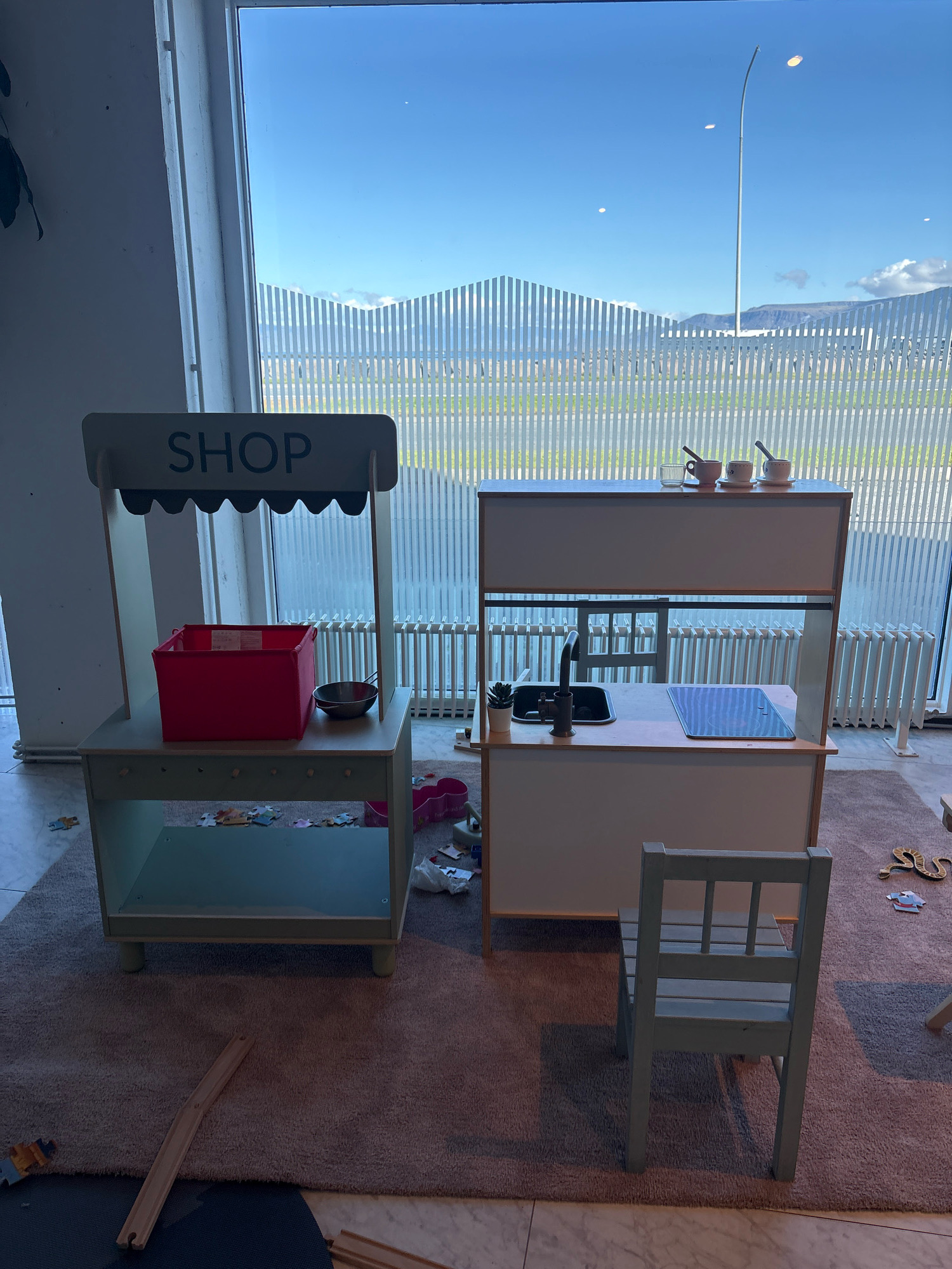 This is a medium shot of a children's playroom with two play kitchen sets in front of a large window overlooking a cityscape and sky in Reykjavik, Iceland. The left play set resembles a shop with a sign reading "SHOP" and a red fabric bin. The right play set is a kitchen unit with a sink, faucet, and stovetop. A small wooden chair sits in front of the kitchen set. Scattered on the floor are puzzle pieces and other toys. The window reveals a bright blue sky with some clouds, mountains in the distance, and a unique fence-like structure. The overall scene suggests a bright, modern, and child-friendly indoor space.