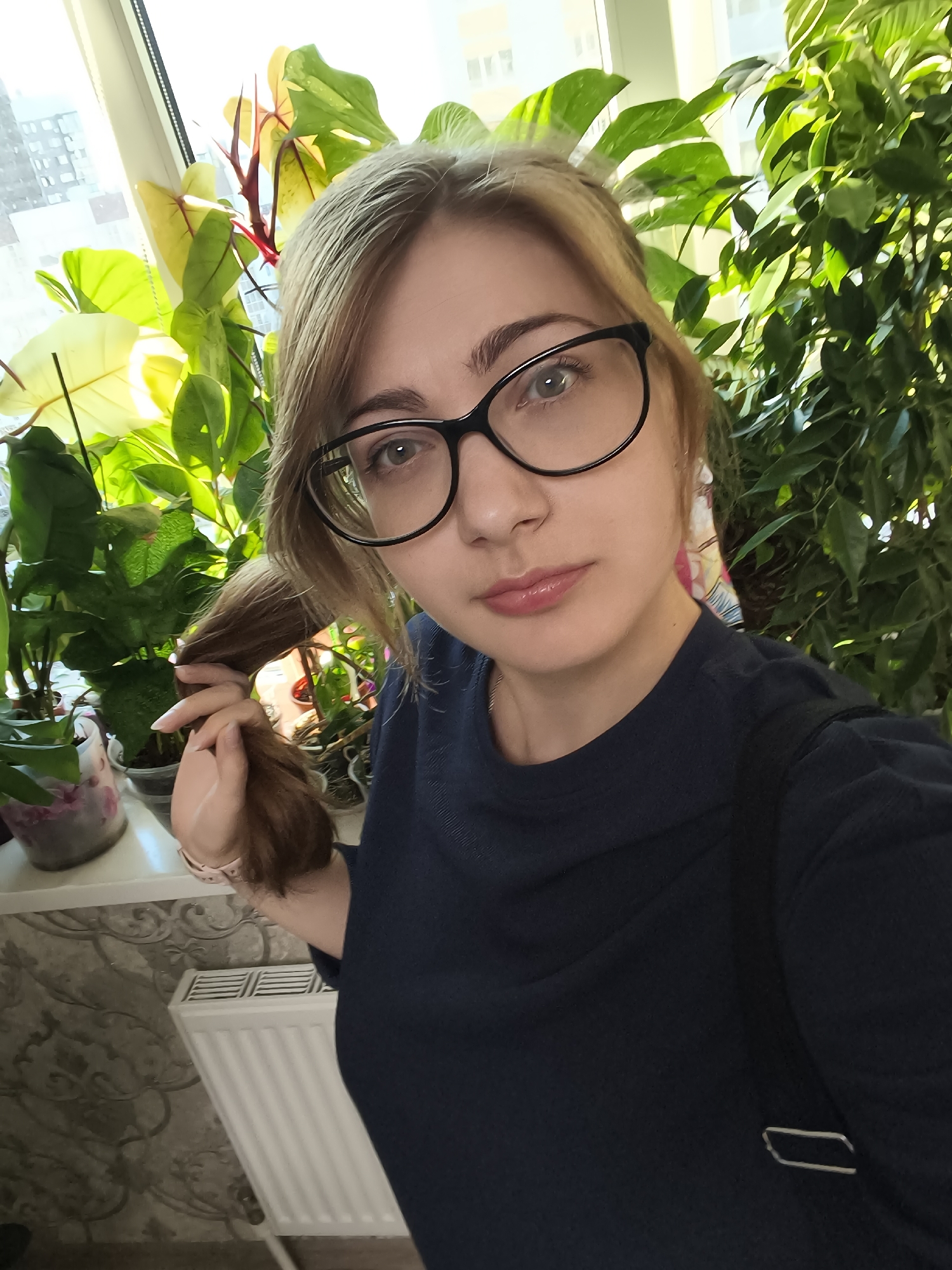 A woman with blonde hair and glasses is taking a selfie in front of houseplants. She is wearing a dark blue shirt and has her hair pulled to the side, holding a strand of it with her hand. The background features a window with natural light, several large green plants, and a radiator. The setting appears to be indoors, possibly a home, with sunlight streaming through the window, suggesting it is daytime. The woman's expression is neutral.