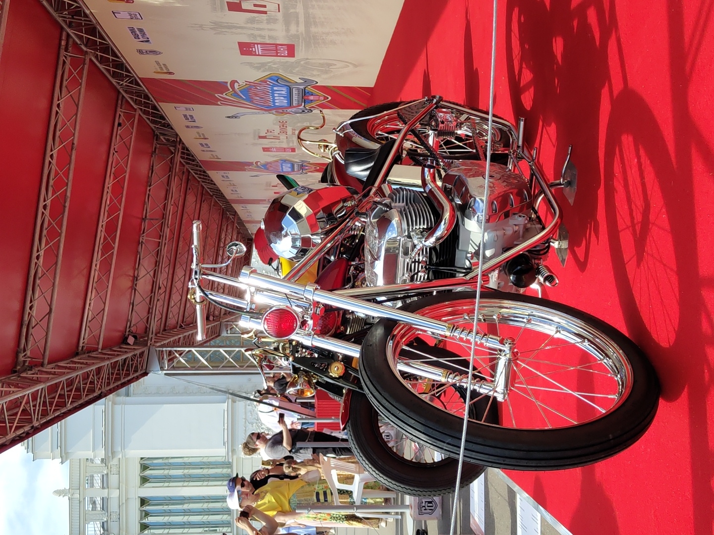An eye-level, medium shot shows a custom red and chrome motorcycle on display at an outdoor event. The motorcycle is the primary subject, positioned prominently in the foreground. Its chrome engine, handlebars, and spoke wheels gleam under the bright sunlight. The motorcycle is set against a vibrant red carpet, which also features a prominent shadow cast by the vehicle. In the background, a red canopy structure with metal supports stretches across the scene, suggesting an event setup. Behind the canopy, a white building with arched windows is visible, and a banner with Russian text and graphics lines the right side of the frame, indicating the location of the event. A few people, dressed in casual attire, are seen in the background, observing the display. One person in a yellow shirt and white hat is in the left portion of the frame, taking a photo with their phone. The overall environment appears to be a sunny day at a public event in Moscow, Russia, likely a motorcycle exhibition or festival.