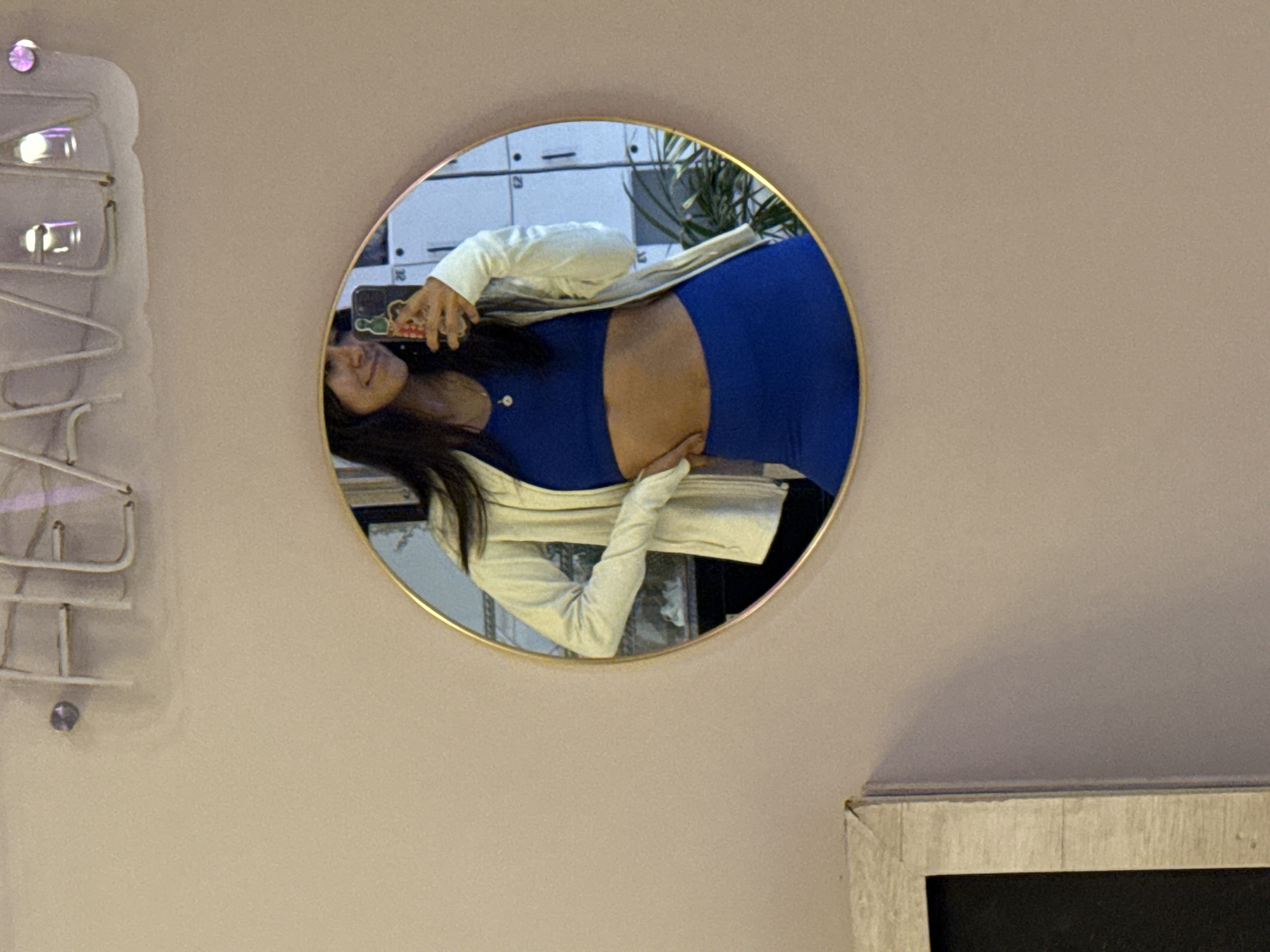The image displays a round mirror with a thin gold frame, mounted on a light beige-pink wall. Reflected within the mirror, a woman is shown horizontally, with her head to the left and feet to the right. She has long dark hair and is smiling faintly, looking towards the camera. She is wearing a royal blue athletic crop top and matching bottoms, partially covered by a light cream or off-white jacket. Her right hand is gently placed on her exposed belly, suggesting she is pregnant. She is holding a smartphone with a distinctive green cartoon-character phone case, using it to capture the mirror selfie. The number '32' is faintly visible on the phone case.
The background reflected in the mirror behind the woman includes white locker-like units and a green potted plant, indicating an indoor setting possibly a gym, studio, or recreational facility. Outside the mirror, to the left, a white metal rack or decorative element with glowing light fixtures is mounted on the wall. In the bottom right corner of the image, a portion of a framed item with a rustic light-colored wooden frame and a dark interior is visible. The scene is contextually located in Puebla, Mexico.