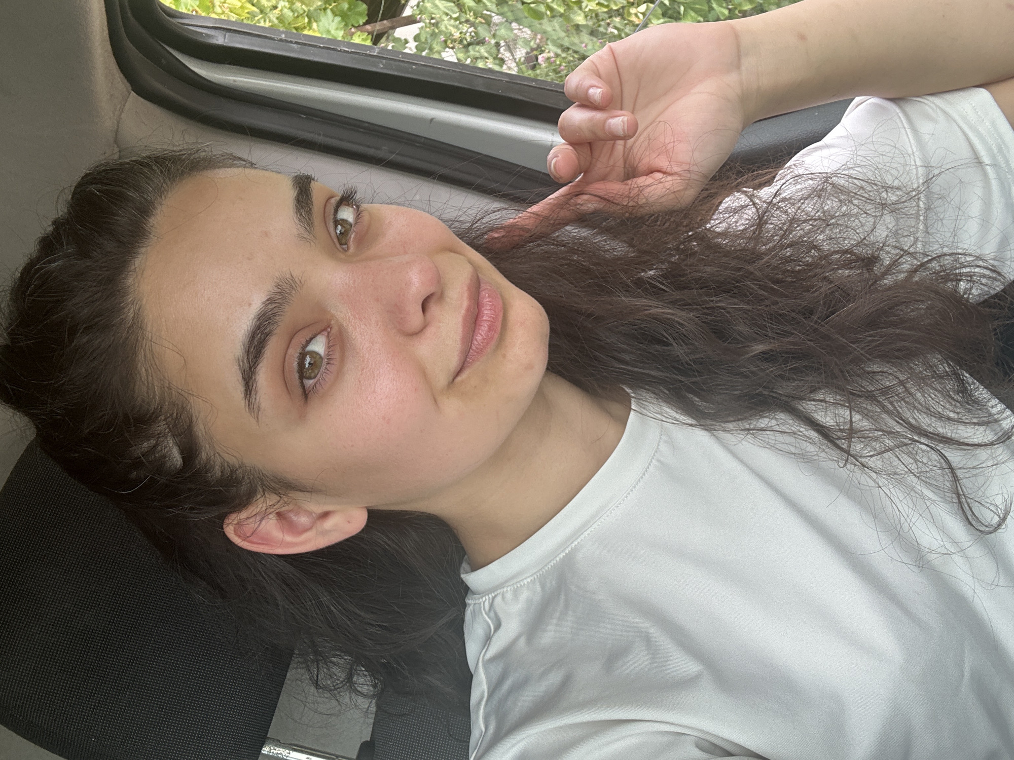 A young woman with dark, curly hair and brown eyes is captured in a selfie taken inside a vehicle. She is wearing a white t-shirt and appears to be holding a strand of her hair near her face. The background shows the interior of the vehicle, including a dark headrest and a window with greenery visible outside. The image suggests an outdoor setting, possibly on a road or near a natural area within or near the city of Malevo, Bulgaria.
