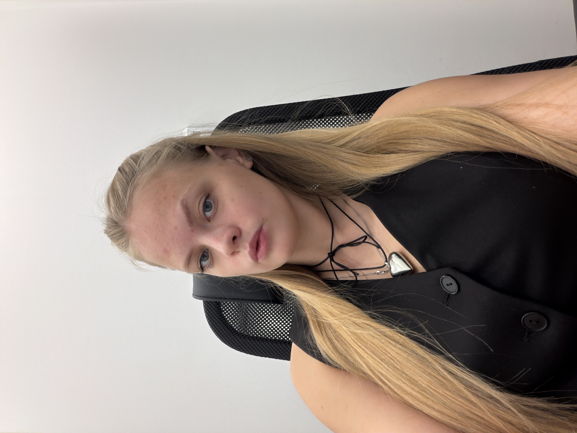 A woman with long blonde hair sits in a black office chair against a white wall. She wears a black sleeveless top with two visible buttons and a necklace with a heart-shaped pendant. The location is not identifiable as Izhevsk, Russia.