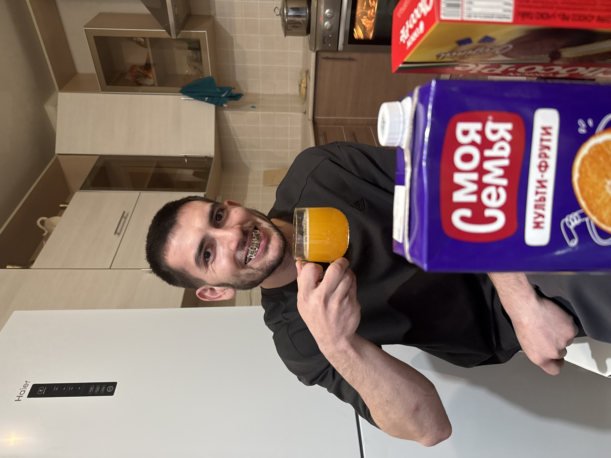 A man with short dark hair and braces is holding a glass of orange juice in his right hand. He is wearing a black t-shirt. A carton of "Moya Semia" (My Family) multi-fruit juice is in the foreground. Behind him is a white Haier refrigerator. In the background is a kitchen with light-colored cabinets and an oven. Based on the packaging of the juice, this photo was likely taken in Russia. Given the appearance of the kitchen and the man, it is likely in an apartment in the city of Kazan, Russia.
