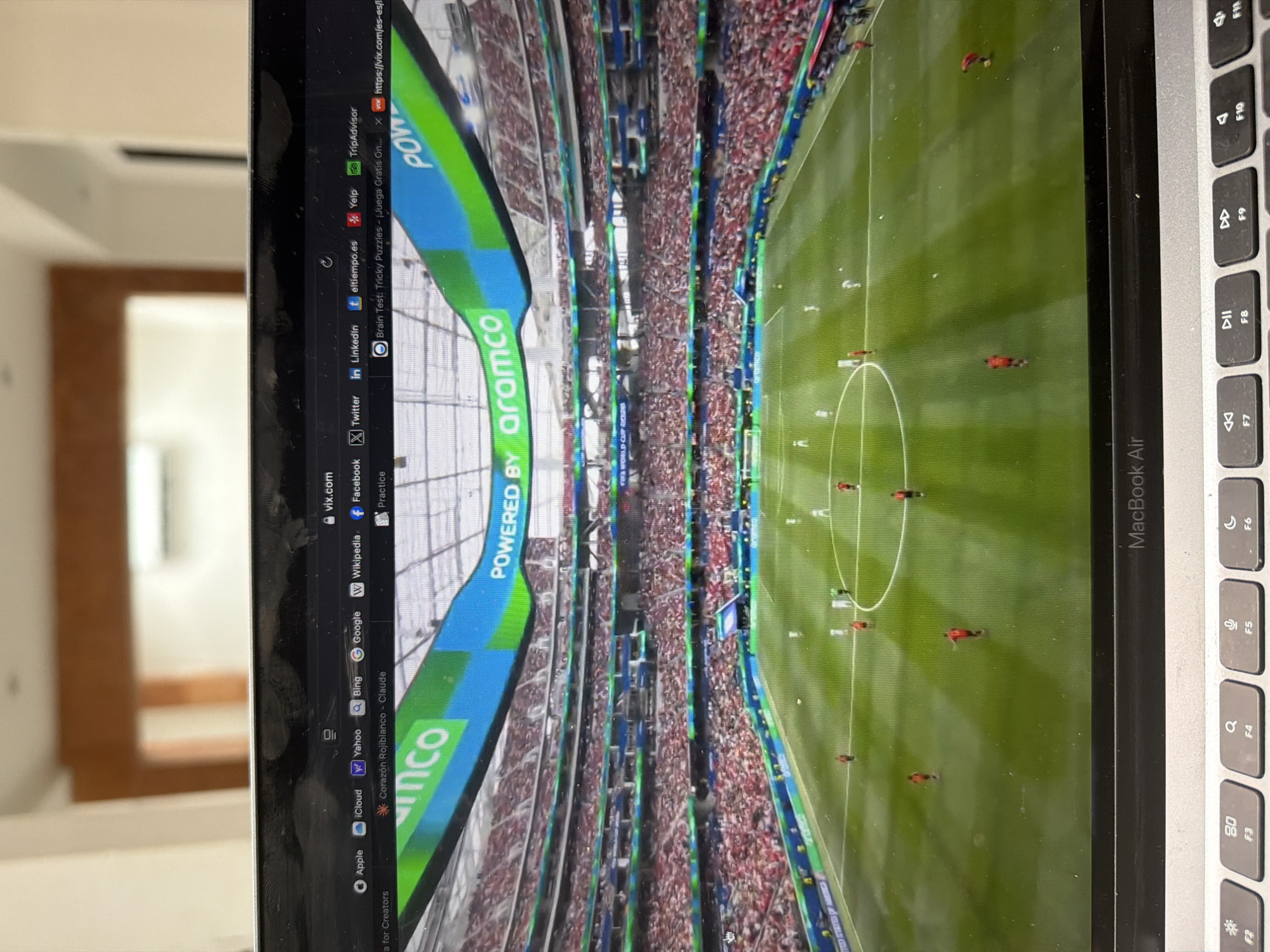 The image shows a laptop screen displaying a soccer game in a stadium. The stadium is filled with spectators wearing red attire. The screen also shows various browser tabs, including "vix.com," "eltiempo.es," and "TripAdvisor." The laptop itself is placed on a keyboard. The background of the image is blurry but suggests an indoor setting with wooden furniture and lighting. The location is not specified in the image or the surrounding context as Cancún, Mexico.
