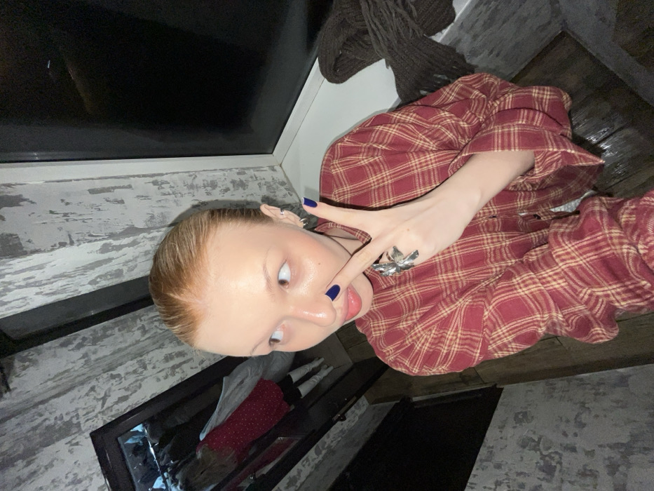 An individual, appearing female with fair skin and red-blonde hair pulled back, is depicted indoors. The subject wears a red and yellow plaid long-sleeved shirt. The left hand displays dark blue nail polish on the fingernails, a large metallic ring on the index finger, and is posed in a "peace" sign gesture with the index finger touching the nose. The individual's tongue is slightly extended, and multiple small hoop earrings are visible on the left ear. The background features a textured wall with a distressed light grey and white finish. A dark-framed mirror on the left reflects another individual, possibly the same person, wearing a red polka-dot dress and white boots. To the right, a dark-framed window or panel is visible, with a dark brown knitted item resting on the sill beneath it. The floor appears to be dark wood or laminate. The specific location within the city of Chaikovsky, Russia, cannot be determined from the image.