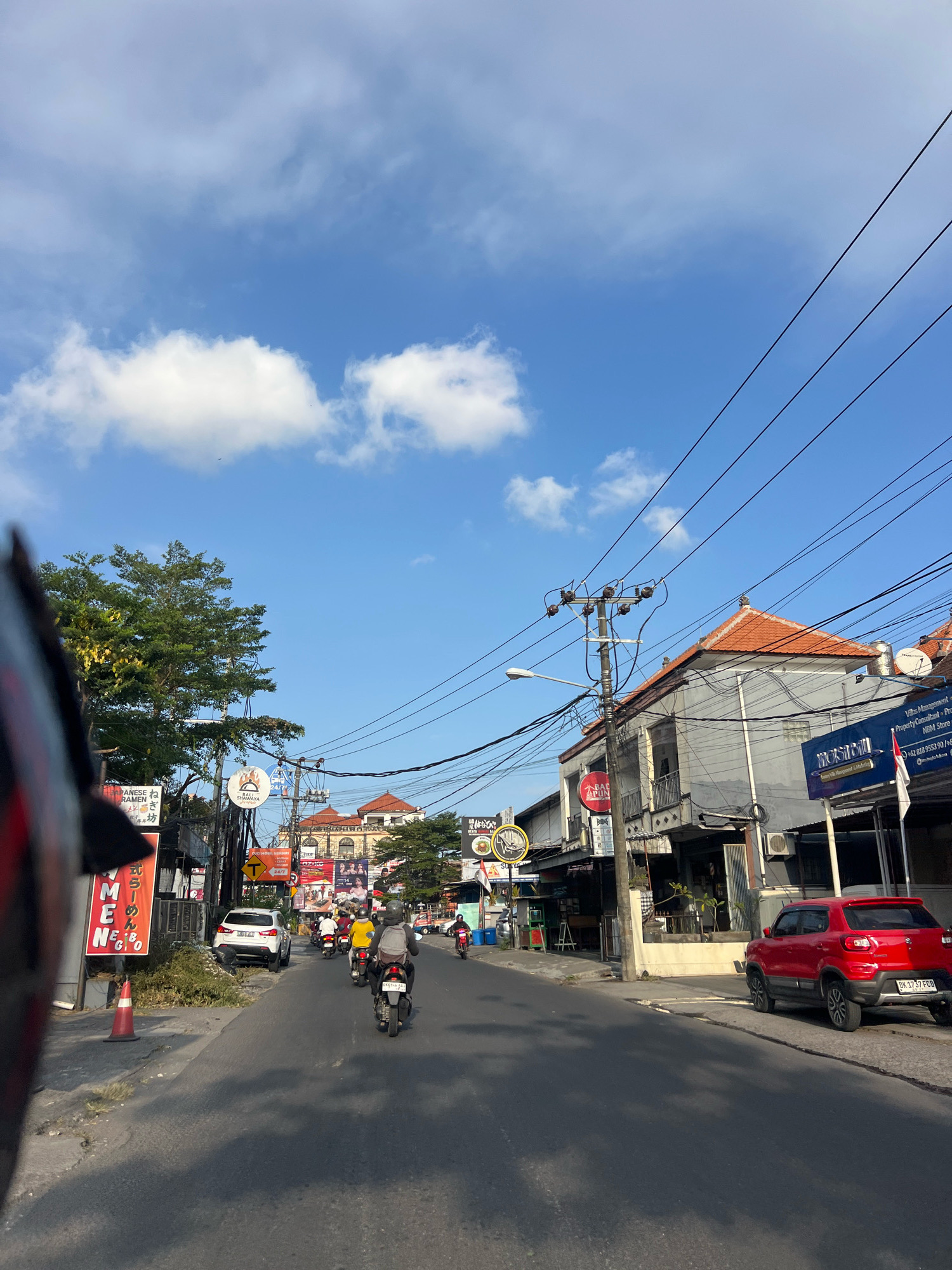 The image captures a street scene in Denpasar, Indonesia, under a bright blue sky with scattered clouds. The perspective suggests the photo was taken from a moving vehicle, possibly a motorcycle, due to the blurred helmet visor in the foreground. The street is lined with shops and buildings, many with signage in what appears to be Indonesian or a similar script.
Several motorcycles and a white SUV are present on the road. One person on a motorcycle wears a yellow shirt and another a gray backpack. The architecture features tiled roofs, common in tropical regions. Power lines are prominently strung across the sky, supported by utility poles. A red car is parked on the right side of the street. The general atmosphere is that of a busy urban environment during daylight.