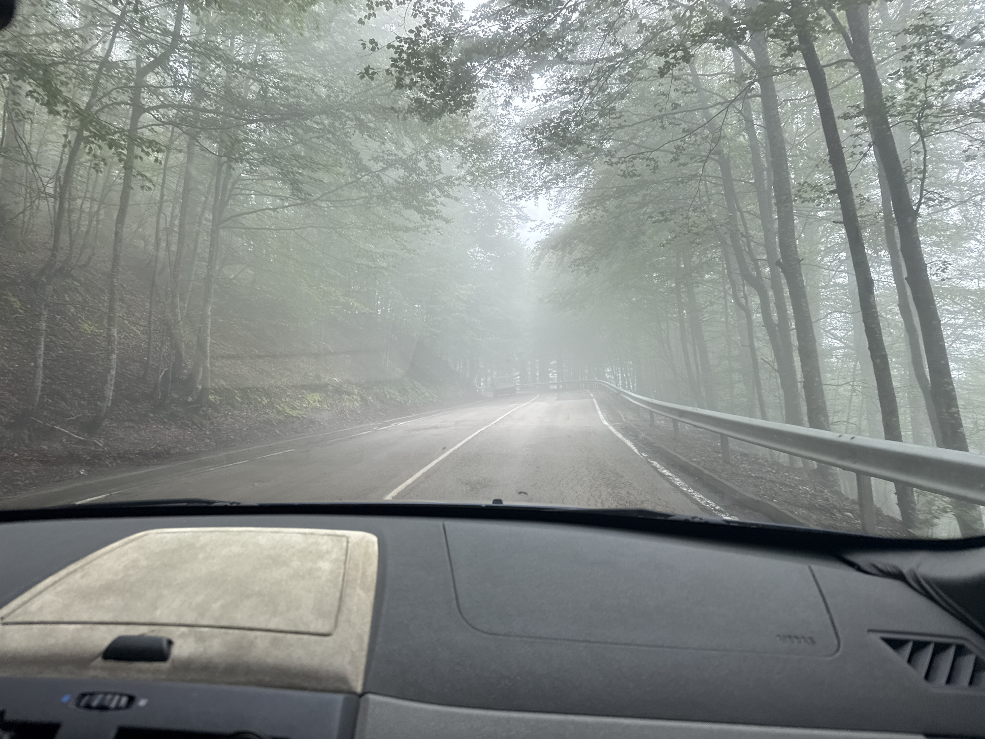 The image is taken from the interior of a vehicle driving on a road through a foggy forest. The road is paved and has a white line marking the lanes. On the right side of the road, there is a metal guardrail. The forest consists of tall trees with dense foliage, and the fog creates a hazy, ethereal atmosphere. The trees are predominantly deciduous, with green leaves visible through the mist. In the foreground, the dashboard of the vehicle is visible, with a textured panel and a small black object. The lighting is dim and diffused due to the fog, creating a moody and atmospheric scene. The location is specified as Beli Osam, Bulgaria. There are no people or animals visible in the image. The overall impression is one of a quiet, serene drive through a misty woodland.