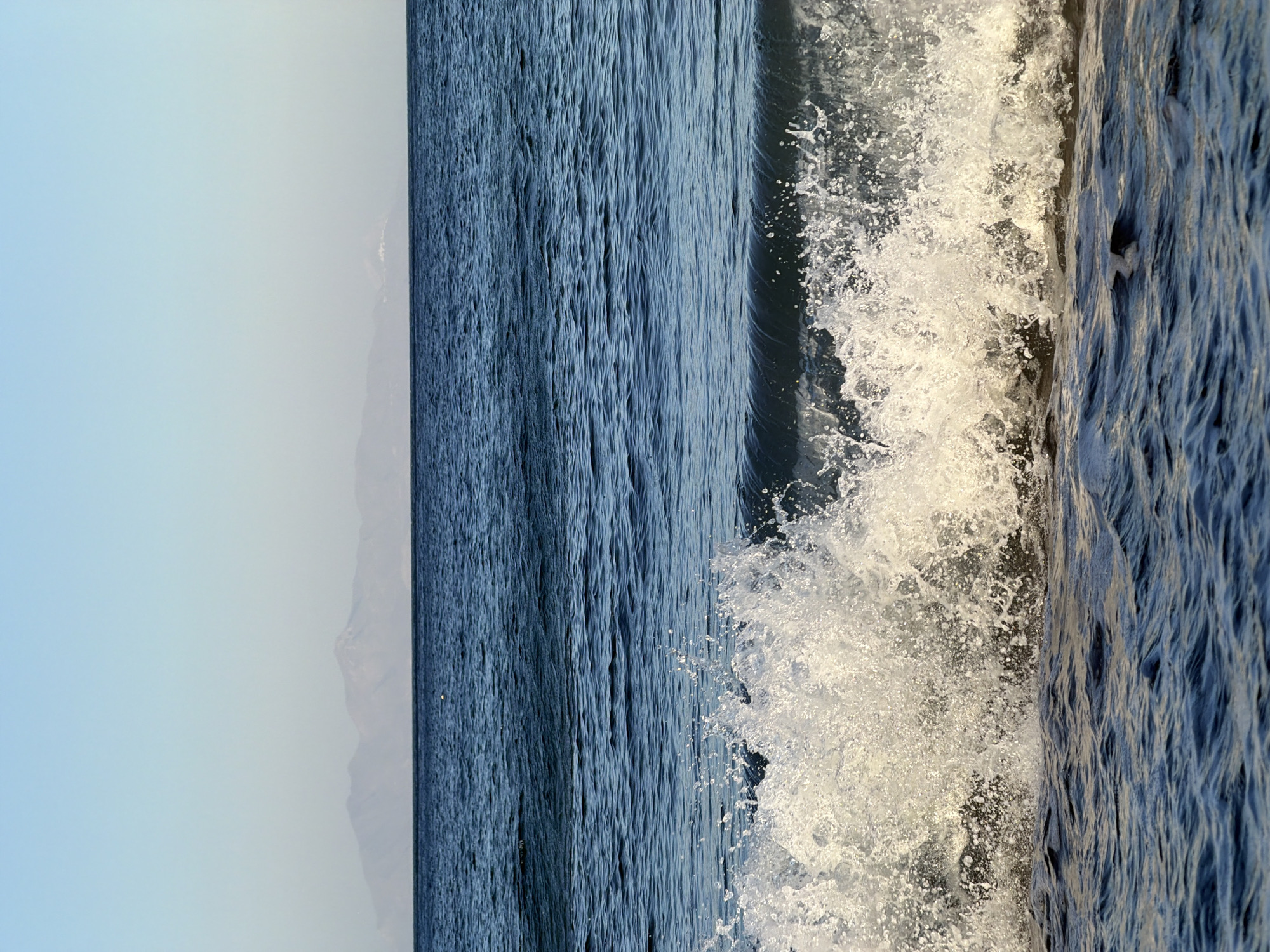 The image shows a seascape with a breaking wave in the foreground and a distant mountain range on the horizon. The water is a deep blue, and the sky is a pale blue with a slight haze. The wave is captured in mid-break, with white foam and spray erupting upwards. The distant mountains appear hazy and are silhouetted against the sky. There is no indication of a specific location within Thira Municipal Unit, Greece, as no landmarks or identifying features are visible. The scene is devoid of people or any objects other than natural elements.