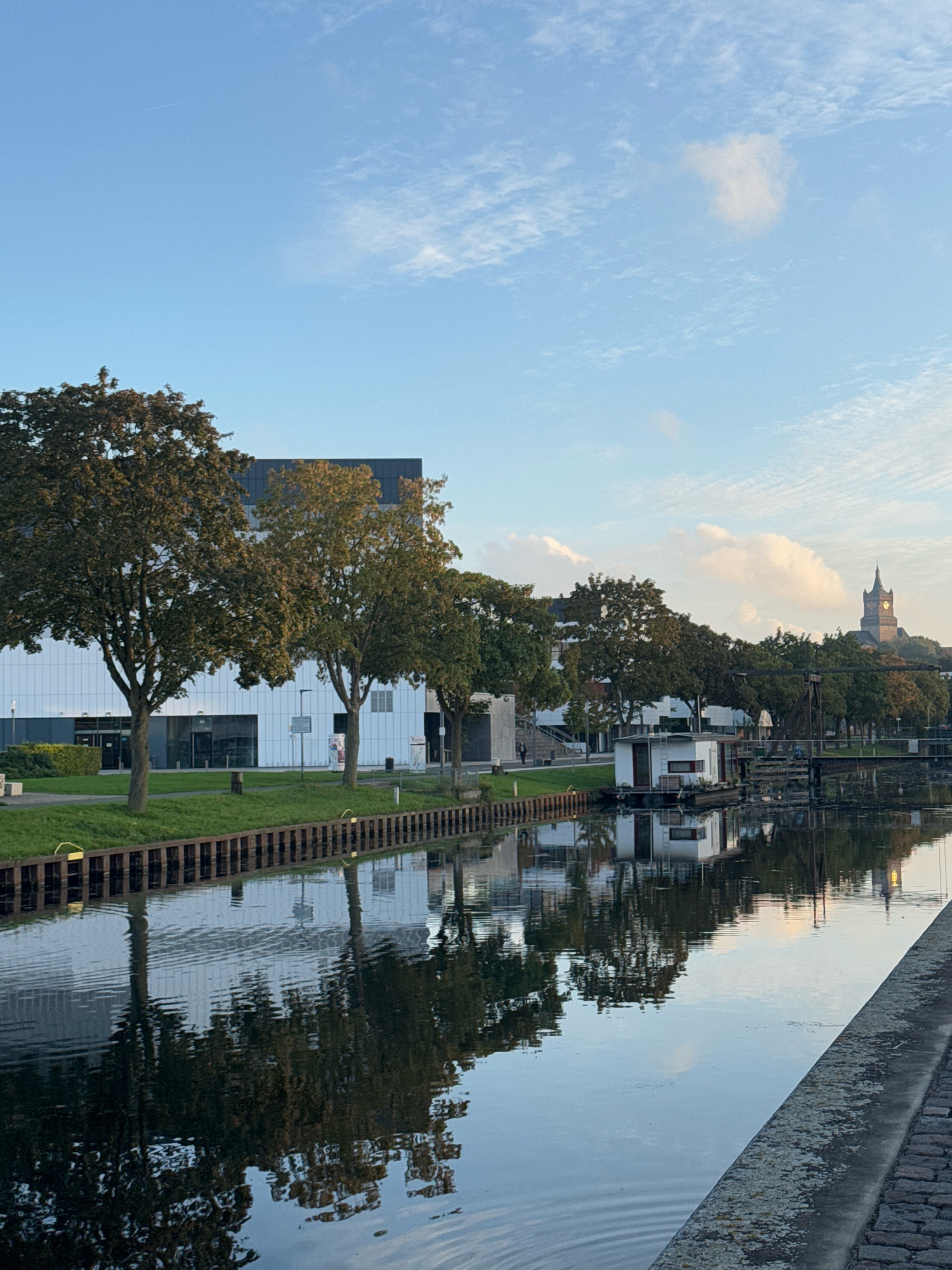 This image depicts a canal in the city of Cleves, Germany, as seen from a cobblestone embankment. On the left side of the canal, a modern building with a white facade and dark roof is visible, fronted by several large trees with green and yellowing leaves. A narrow strip of grass separates the building from the canal, which is bordered by a wooden retaining wall with yellow bollards. The calm water of the canal reflects the building, trees, and sky.
In the middle of the canal, a white houseboat with red trim is moored. Beyond the houseboat, more trees line the far bank, and a dark, industrial-looking structure with a bridge mechanism is present. In the distance, a church tower with a pointed roof and illuminated clock face rises above the trees, suggesting the presence of a historical district. The sky is a light blue with scattered white clouds, and a faint contrail is visible in the upper left corner. The lighting suggests it is either early morning or late afternoon. There are no discernible people or animals in the image.