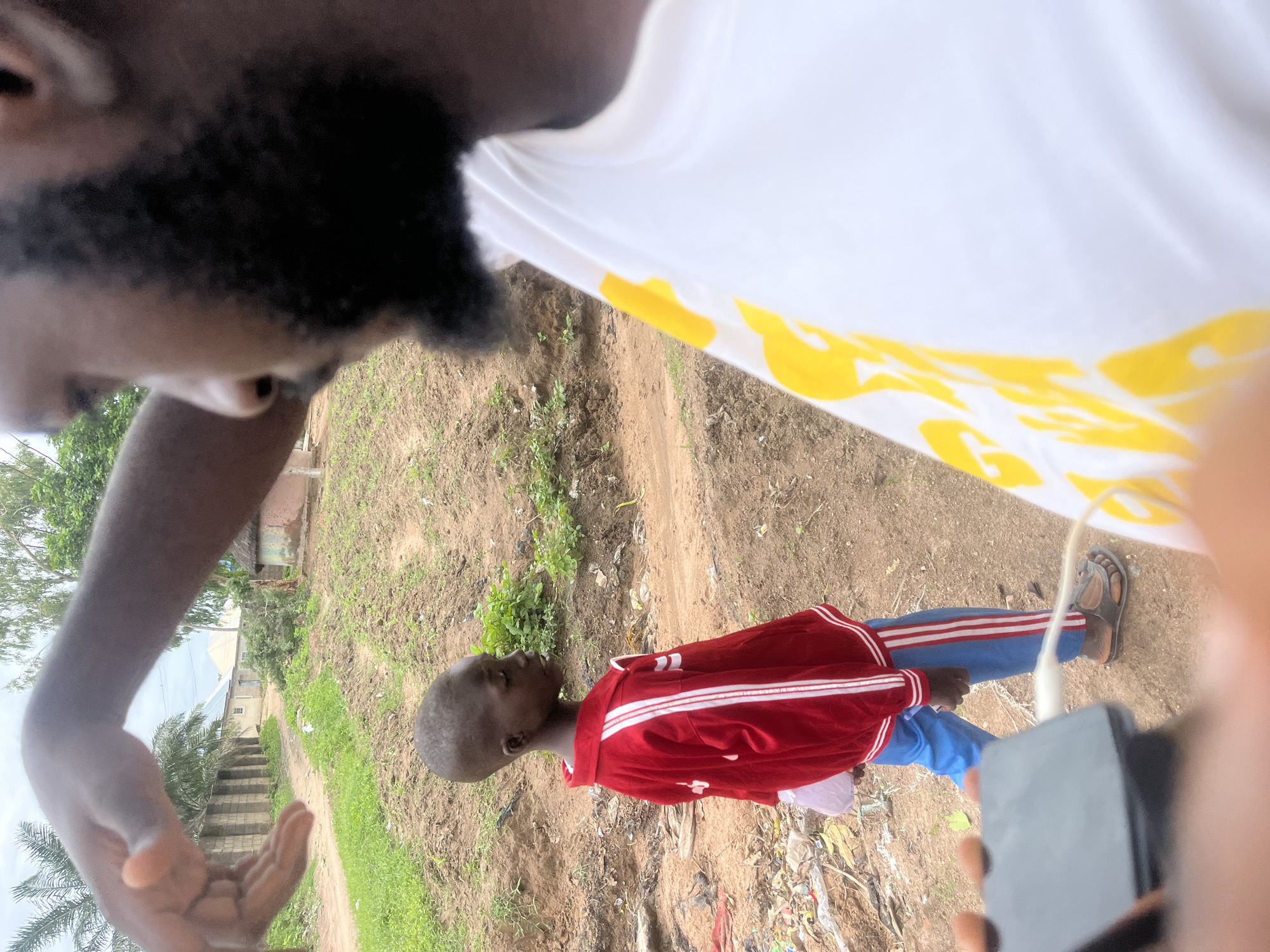 In Jalingo, Nigeria, a young boy, appearing to be around eight years old, stands looking to his right, wearing a red jacket with white stripes on the sleeves and collar, and blue athletic pants with red and white stripes. He is also wearing brown sandals. The boy is in a dirt area with sparse vegetation, and behind him are some buildings and a fence. In the foreground, an adult's arm and hand are visible, and a white shirt with yellow designs is worn by the adult. A dark-colored smartphone is held in the foreground, possibly recording the scene. The overall environment suggests a rural or semi-urban setting.