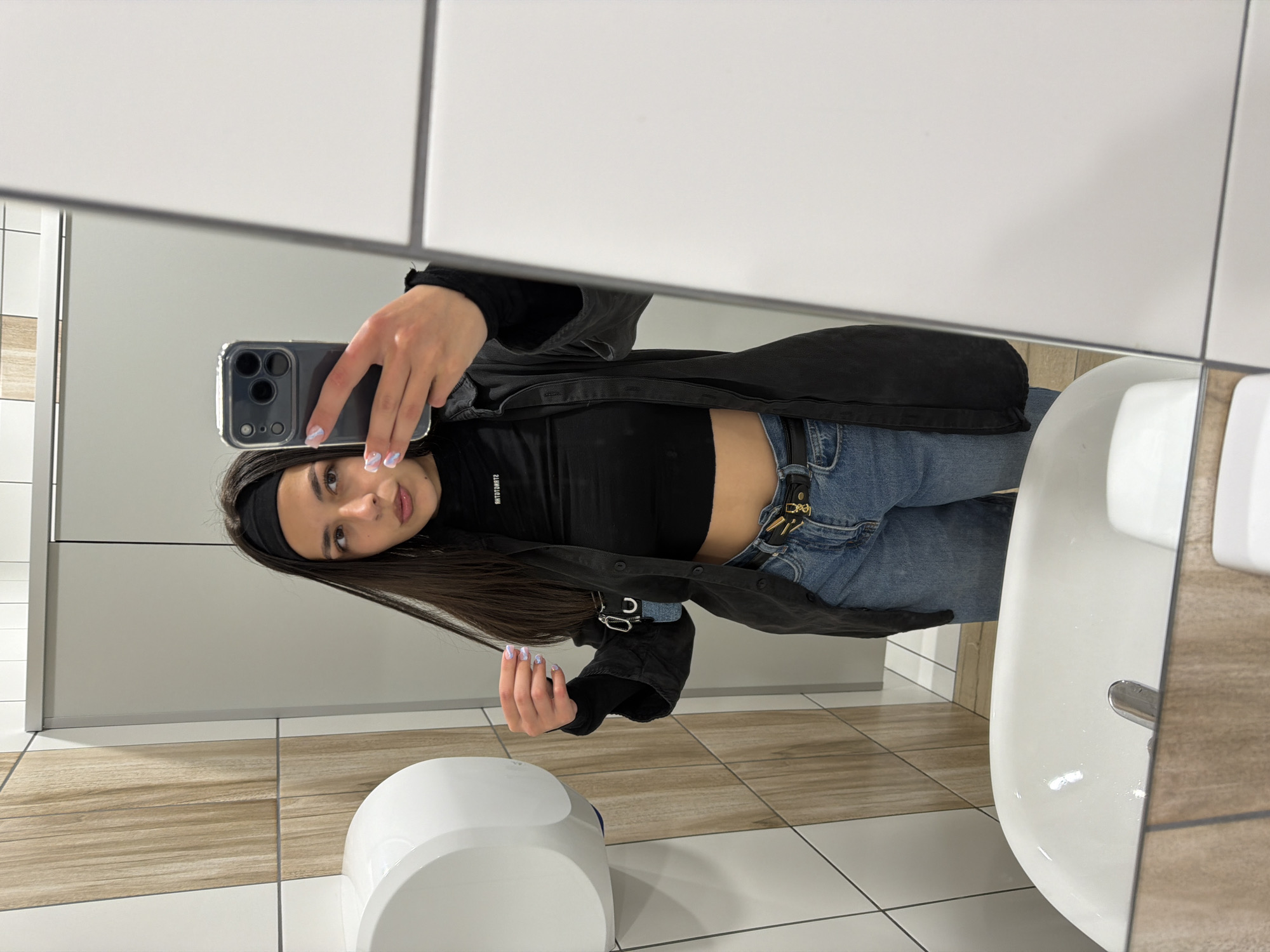 A young woman is taking a selfie in a public restroom. The restroom has white and beige tile walls and flooring. A white automatic hand dryer is visible on the left, and a white sink is on the right. The woman is wearing a black top, jeans, and a black jacket. She is holding a smartphone with a clear case. The photograph does not provide enough information to determine the location in the city of Sofia, Bulgaria.