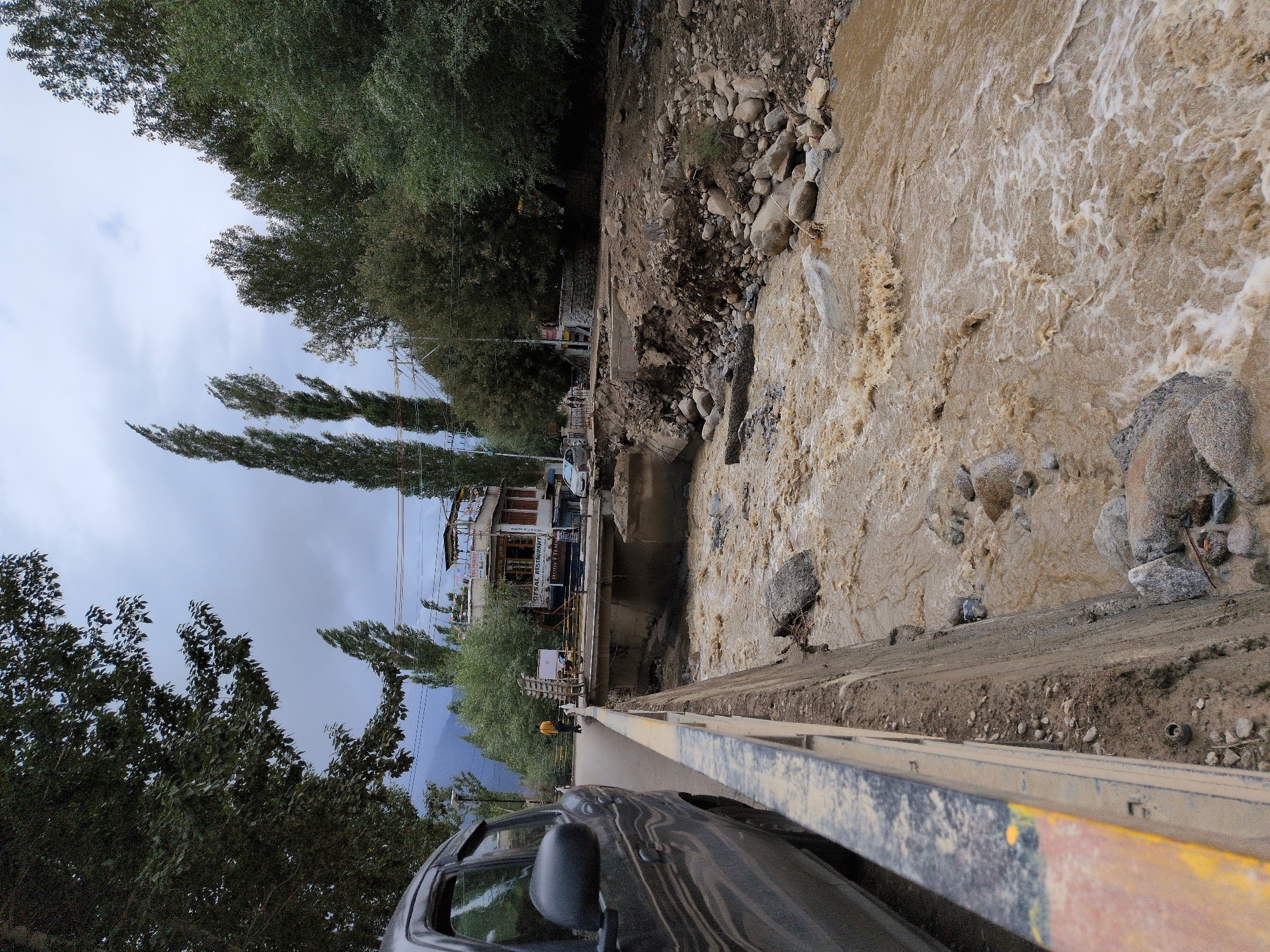 This is a forensic, matter-of-fact description of the image:
The image depicts a scene of a flash flood in Leh, India. A car is parked on the side of a road next to a concrete barrier. The road itself appears to be elevated above a muddy, fast-flowing river. The river is swollen and carries debris, with visible turbulence and white water. The riverbank on the right side of the image shows signs of erosion, with collapsed earth and exposed rocks.
In the background, a two-story building with balconies is visible, along with trees and mountains in the distance. A person in a yellow shirt is standing on the road, observing the flooded river. Electrical wires and poles are present in the background, indicating infrastructure in the area. The sky is overcast, suggesting potentially inclement weather conditions that may have contributed to the flood. The overall scene suggests significant water damage and disruption due to the natural event.