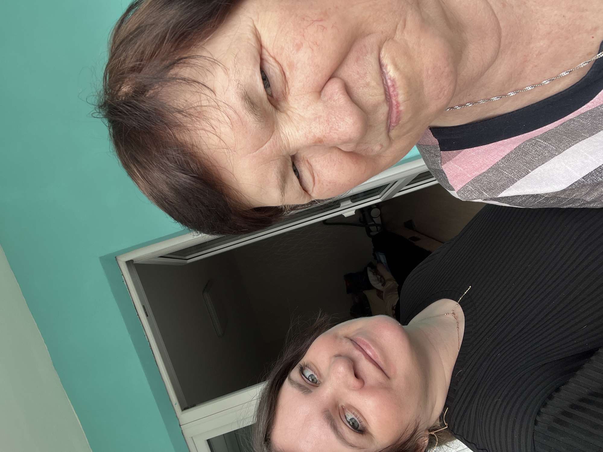Two females are posing for a selfie inside an apartment, the city of Perm, Russia. The first female, an elderly woman with short brown hair and a striped shirt, is positioned towards the top of the frame. The second female, younger with long brown hair and a black top, is at the bottom of the image. A window frame is in the background, along with a turquoise wall. The room appears to be dimly lit.