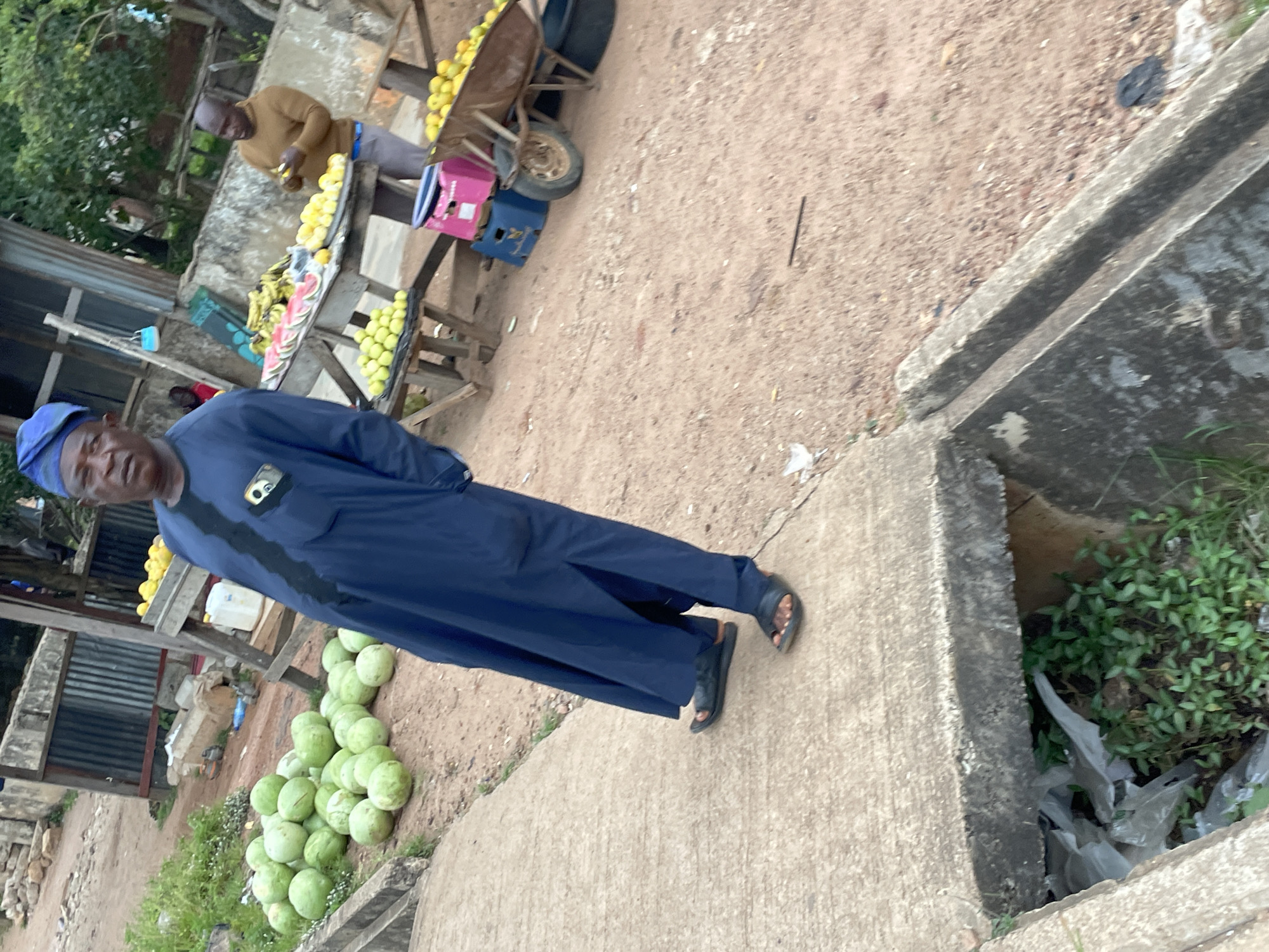 In Jos, Nigeria, an outdoor market scene unfolds. A man, dressed in a dark blue traditional garment and matching cap, stands on a concrete ledge, facing forward. His hands are in his pockets. To his right, a fruit stall displays an assortment of produce. Bananas are visible in bunches, alongside sliced watermelons. Piles of light green, round fruits, possibly small melons or unripe fruits, are stacked on shelves and in a wheelbarrow. Another man, wearing a brown sweater and dark pants, is seen behind the fruit stall, appearing to be handling some of the fruits. The ground is a mix of reddish-brown dirt and scattered debris. In the foreground, a drainage channel is visible, containing some green vegetation and white plastic trash. The background shows a wall with corrugated metal sheeting and some trees.