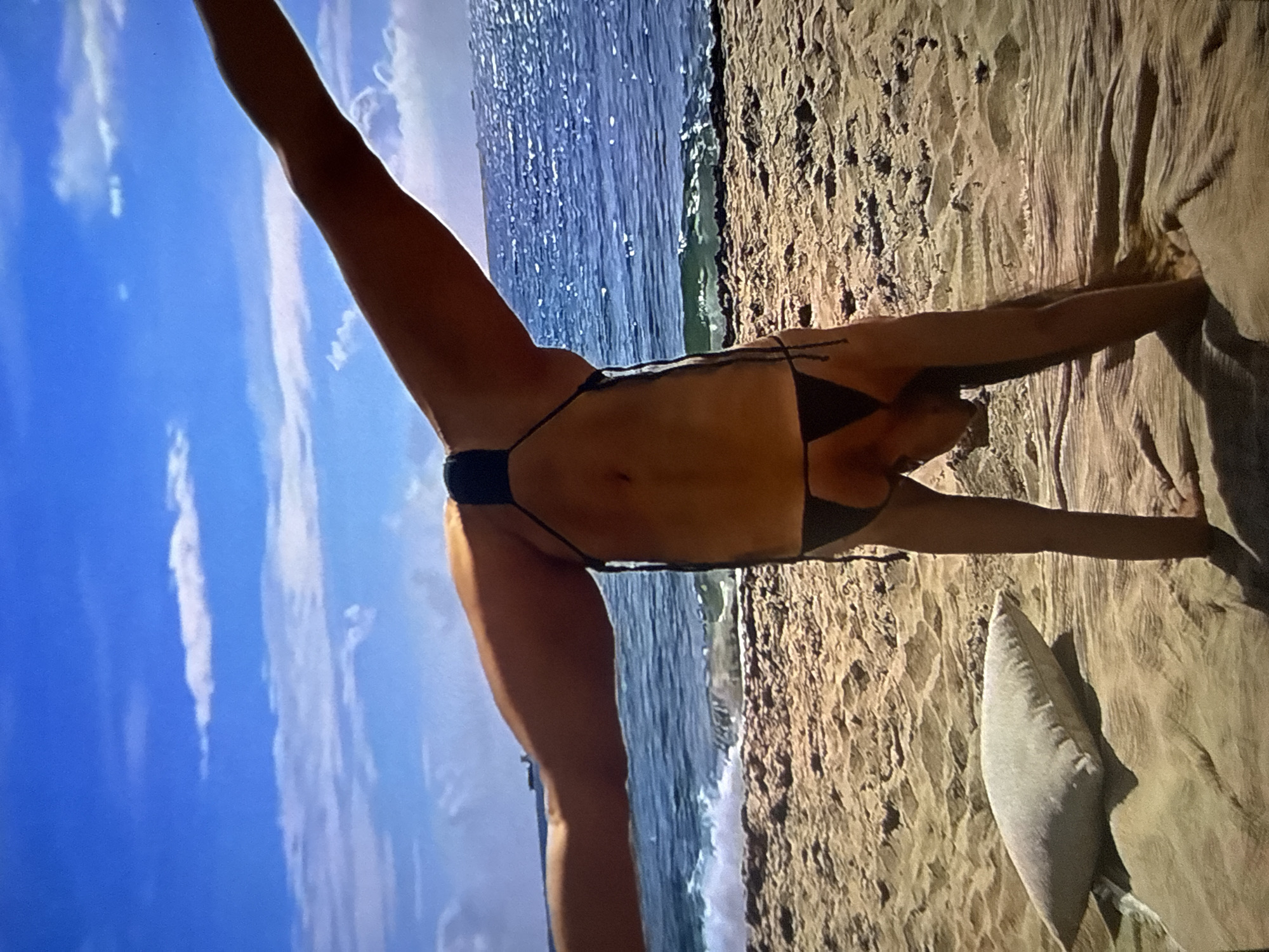 The scene depicts a beach in Varna, Bulgaria. An individual, appearing female, is captured in an inverted position, performing a handstand with her legs extended wide in a horizontal split. She is wearing a dark, minimalist string bikini. Her hands are positioned on a wrinkled, light tan fabric spread on the light brown, textured sand. To her immediate left, a light grey rectangular pillow rests on the sand. In the background, the ocean's dark blue water is visible with small waves breaking at the shoreline, exhibiting numerous specular reflections from bright sunlight. Several large ships are present on the distant horizon line. The sky above is clear blue with scattered white cumulus clouds. The entire scene is illuminated by strong natural daylight.