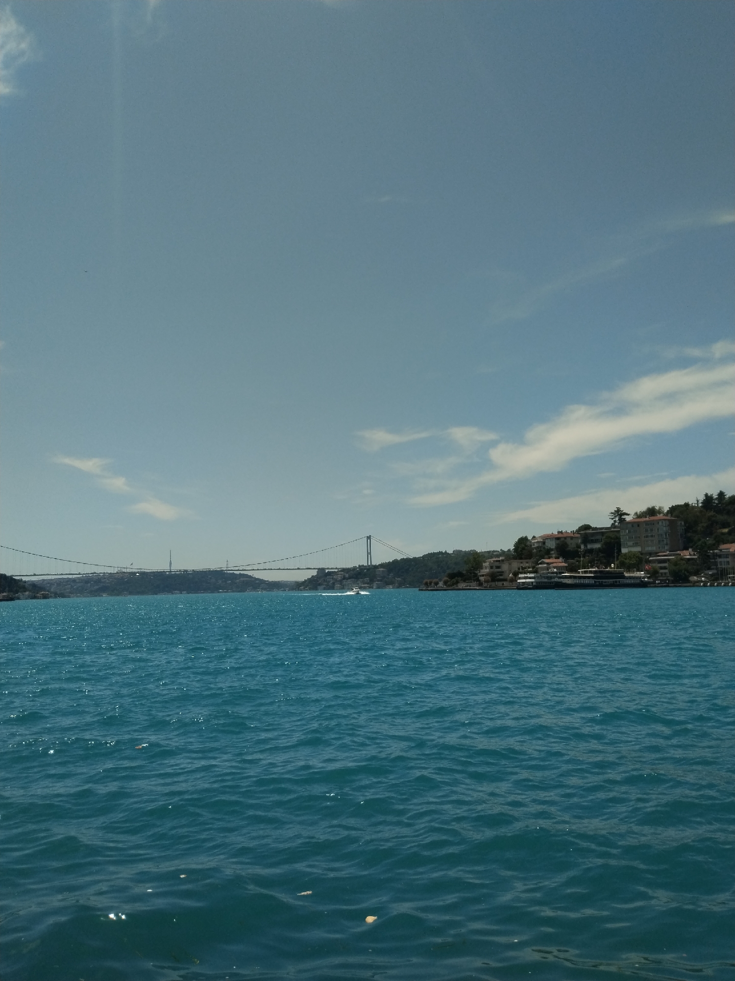 A scenic wide-angle photograph captures a vibrant turquoise-blue body of water under a clear, bright blue sky with scattered white clouds. The sun, high in the sky, casts shimmering reflections on the rippling surface of the water and creates a slight glare in the upper left quadrant.
In the mid-distance, a majestic suspension bridge spans the water, connecting two green, hilly landmasses. The bridge features two tall towers and numerous suspension cables. On the right side of the frame, the lush, green shoreline rises, dotted with various multi-story buildings, likely residences or hotels, nestled among the trees. A long, dark-colored vessel, possibly a ferry or tour boat, is visible near the right bank. Further out on the water to the right, a small white boat can be seen moving, leaving a subtle wake behind it.
The setting appears to be a major strait or waterway, likely in a city known for such bridges and waterfronts, with architectural styles suggestive of the Bosphorus Strait in Istanbul, Turkey. The time of day is clearly daytime, probably mid-day, given the bright illumination and high sun. The weather is sunny and clear. No discernible individuals or text are visible in the image.