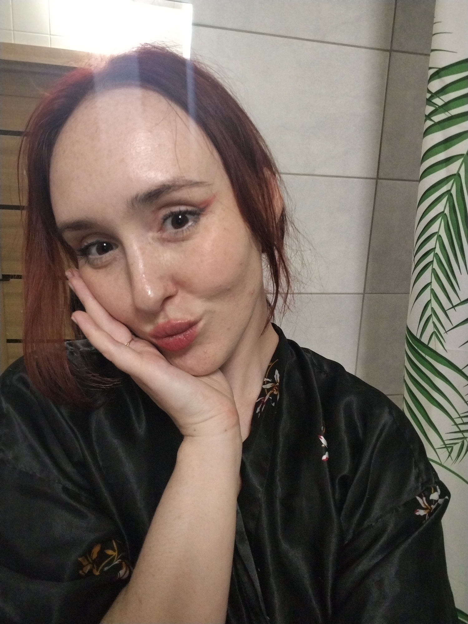 A fair-skinned woman with auburn hair is posing for a selfie indoors. She is wearing a black silky robe with floral accents. Her left hand is raised, with her cheek resting on her palm. She has freckles and is wearing makeup, including red eyeliner. A ring is visible on her finger. The background consists of a white-tiled wall and a shower curtain with a green and white palm leaf pattern. The location is a bathroom. The image does not contain information about the city of Izhevsk, Russia.