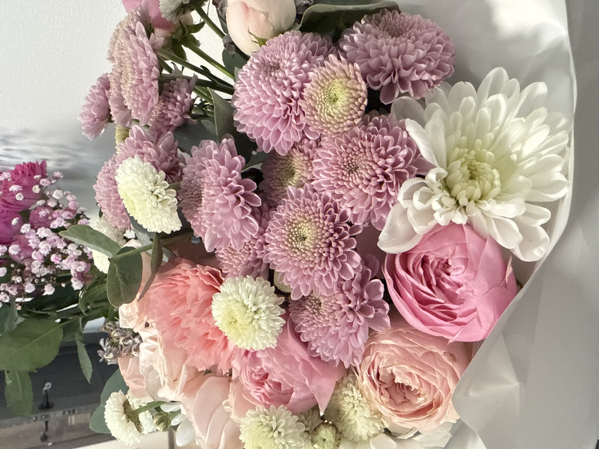 The image shows a bouquet of flowers. The bouquet contains pink and white chrysanthemums, pink roses, and other smaller pink and white flowers. The flowers are wrapped in white paper. The location is likely not in Saint Petersburg, Russia. The image does not provide any information about the city or country.
