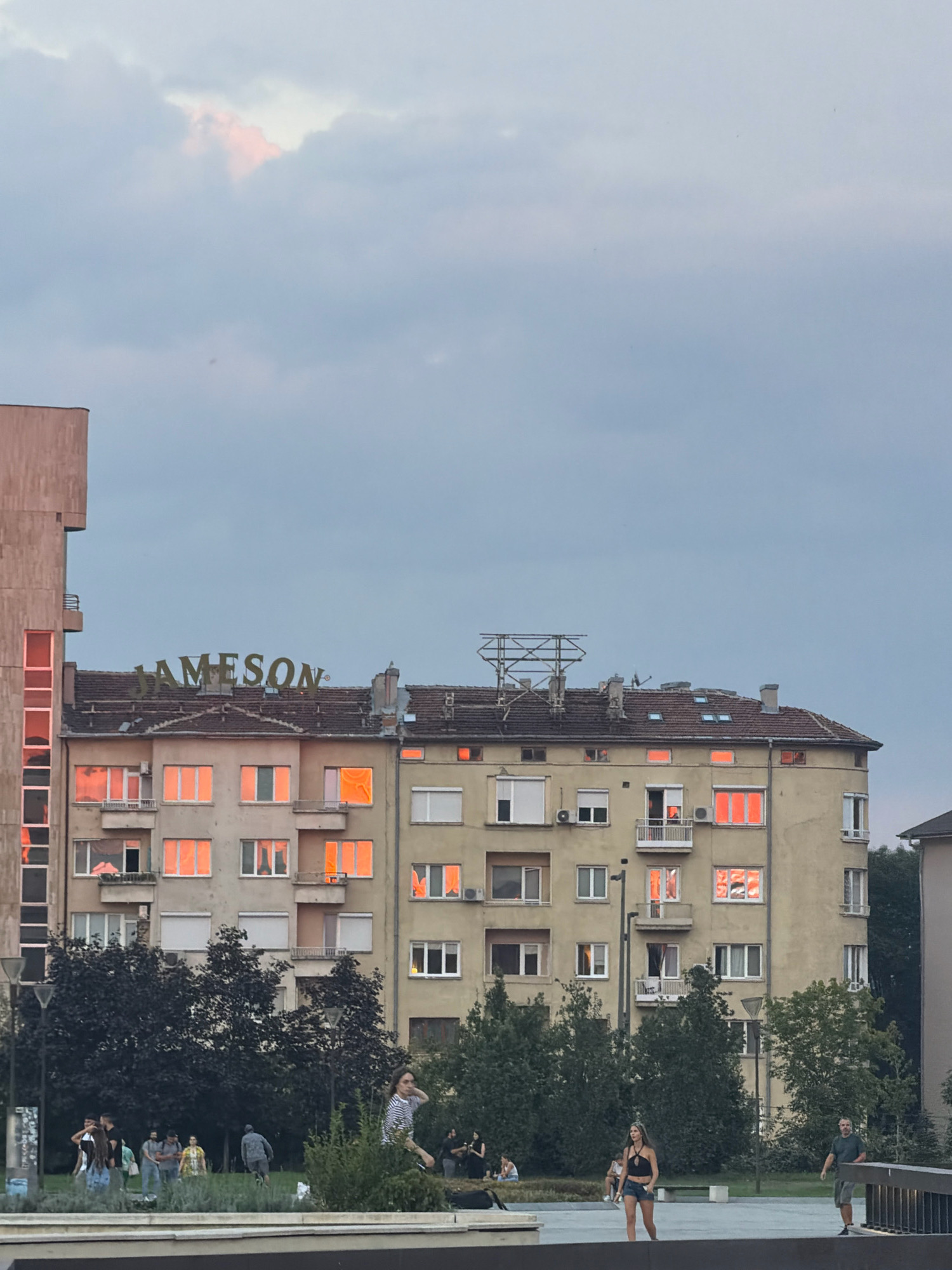 The image depicts a street scene in Sofia, Bulgaria, featuring a multi-story building with numerous windows reflecting a warm, orange light. The building has a large "JAMESON" sign prominently displayed near its roof. Below the building, a park-like area is visible with several trees and a group of people. Some individuals are standing and conversing, while others are seated or walking. A woman in a striped shirt is in motion, and a man in a green shirt and shorts stands near a railing. The sky above is overcast with hints of pink light in the clouds.