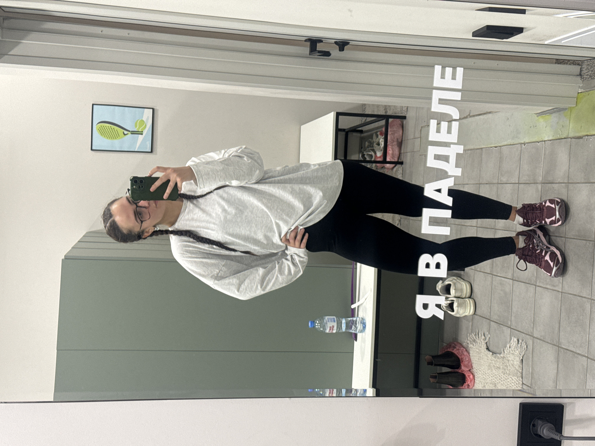 An indoor mirror selfie shows a person with dark braided hair and glasses, wearing a light grey long-sleeved top and black leggings, taking a picture with a dark green smartphone. They are standing upright, with their left hand holding the phone and their right hand resting on their hip. They wear maroon and white athletic shoes.
The setting appears to be a functional indoor space, likely a sports facility or changing area, consistent with the provided location context of Krasnoyarsk, Russia. The floor is tiled grey. Reflected in the mirror on the wall behind the person is artwork depicting a green padel racket and a yellow ball. To the right of the person's reflection, large white Cyrillic letters are visible, spelling "Я В ПАДЕЛЕ" (Ya V PADELE), which translates to "I am in Padel."
Other objects reflected in the mirror include a clear plastic water bottle on a white surface, a pair of white sneakers on the floor, a small white textured rug, and pink slippers. In the background, there's a black metal shelving unit with items inside (possibly shoes or bags) and a light-colored cabinet or wall feature. A black power outlet with a grey plug is visible at the bottom right corner of the mirror's reflection. The time of day is not discernible due to artificial lighting. The person's expression is neutral.