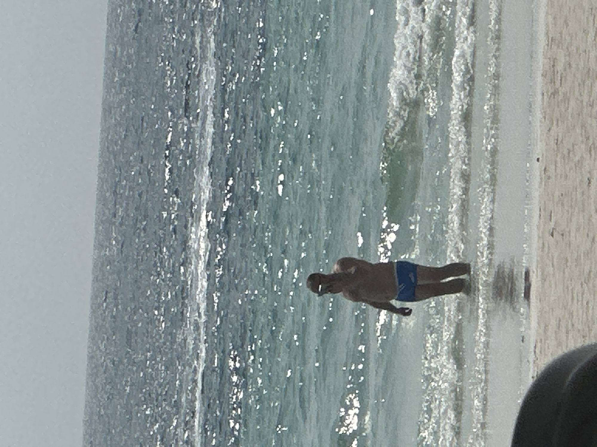 The image depicts a man wearing blue swimming trunks standing in shallow water at a beach in Pattaya City, Thailand. The water is light blue-green with sunlight glinting off the surface. The beach sand is light tan. In the foreground, the lower right corner shows a dark, blurred object. The sky is overcast.
