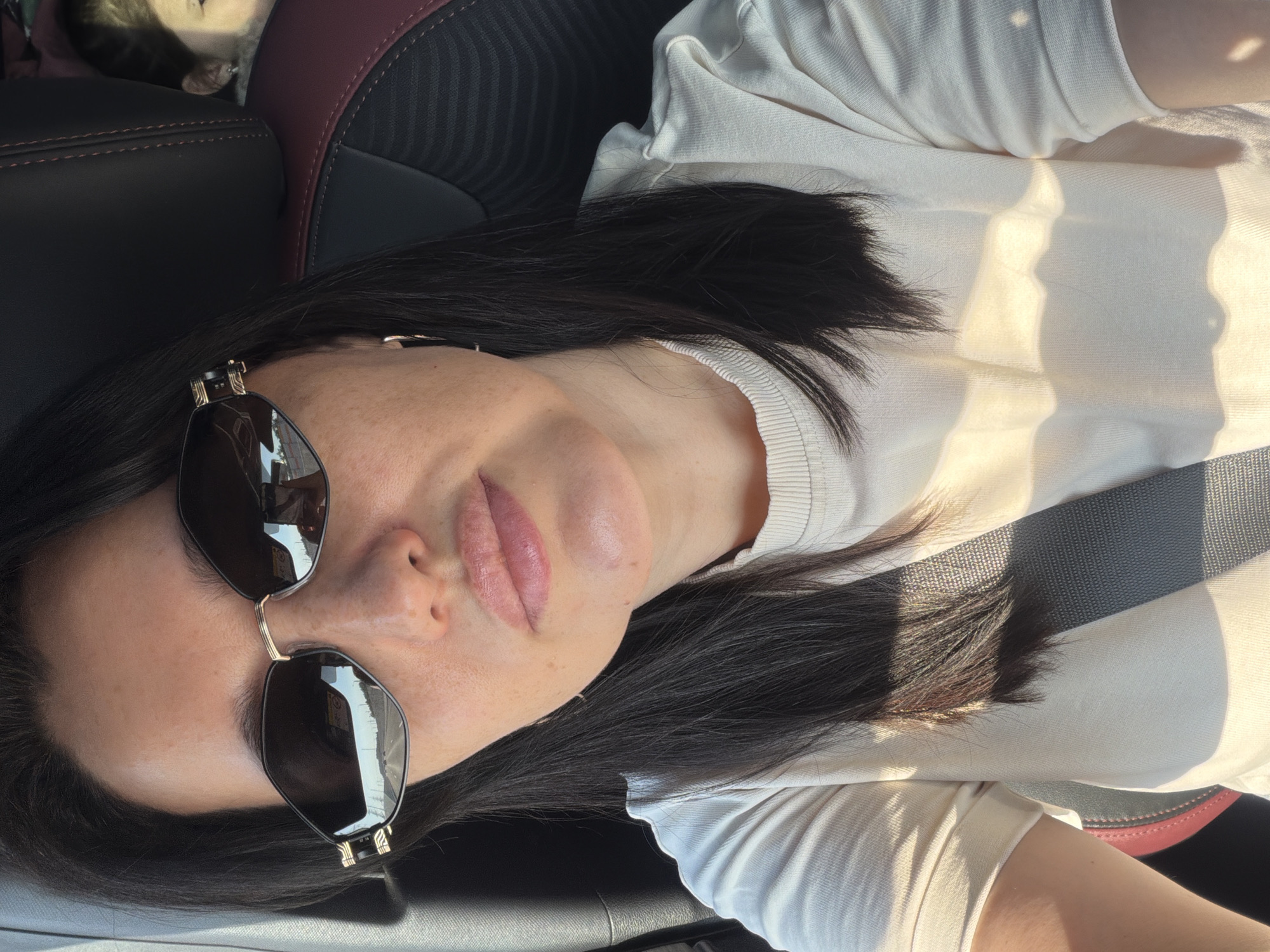 A woman is seen from the chest up, wearing sunglasses and a light-colored t-shirt. She appears to be in the passenger seat of a car, as evidenced by the seatbelt and car interior visible. The woman has dark hair and is looking towards the camera. The background is blurred, but suggests an outdoor environment, possibly a road. The image was taken in Elektrostal, Russia.