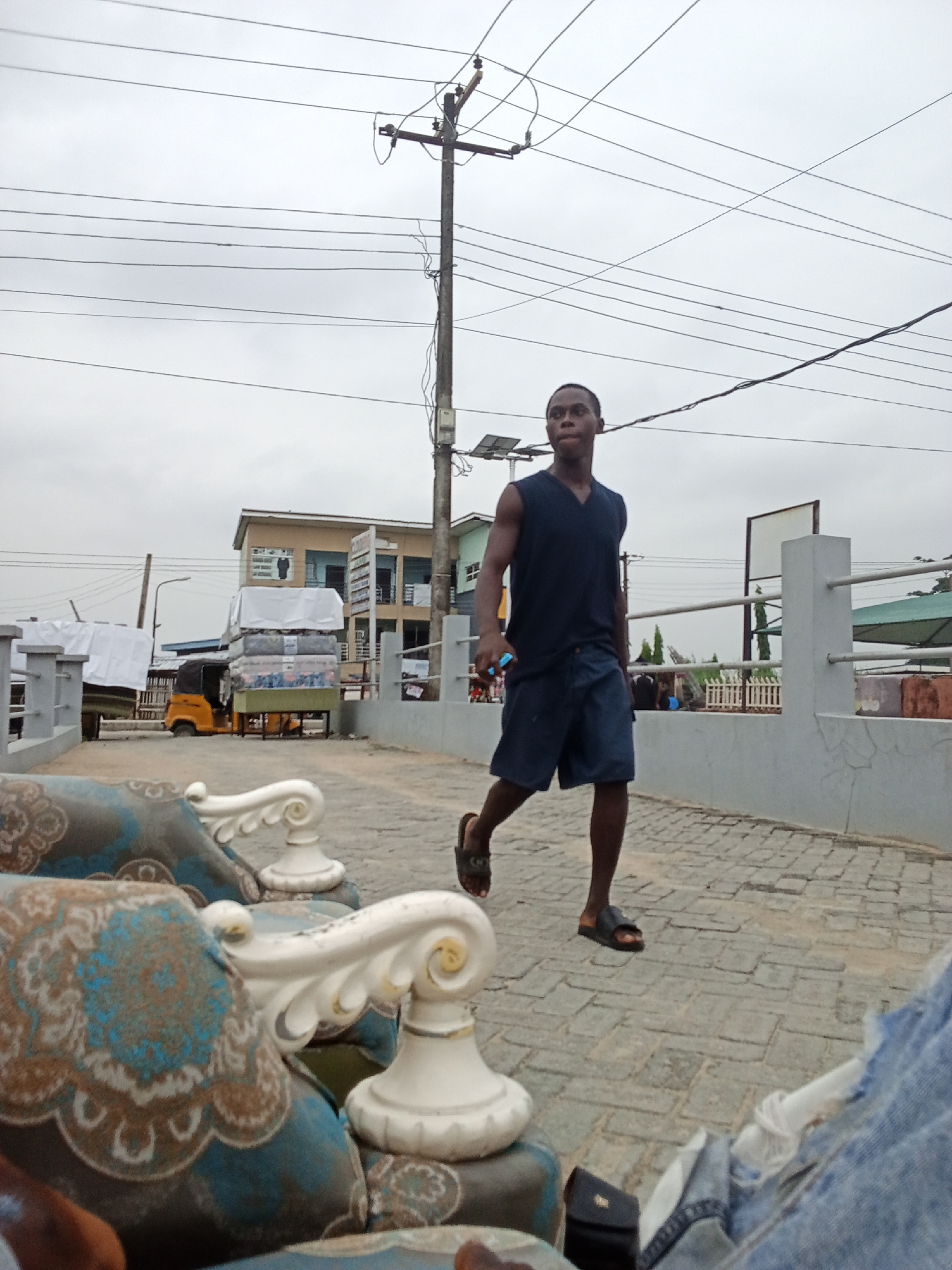 The image depicts a man walking on a brick-paved pathway in Ado, Nigeria. He is wearing a sleeveless navy blue shirt, dark denim shorts, and sandals. He appears to be holding a small, dark-colored object in his right hand. Overhead are numerous power lines. In the background, there is a three-wheeled yellow tricycle loaded with packaged water, situated near a two-story building. In the foreground, parts of a blue-patterned sofa with ornate white armrests are visible, suggesting a vantage point from a seating area. A white concrete barrier runs along the pathway. The sky is overcast, indicating a cloudy day.