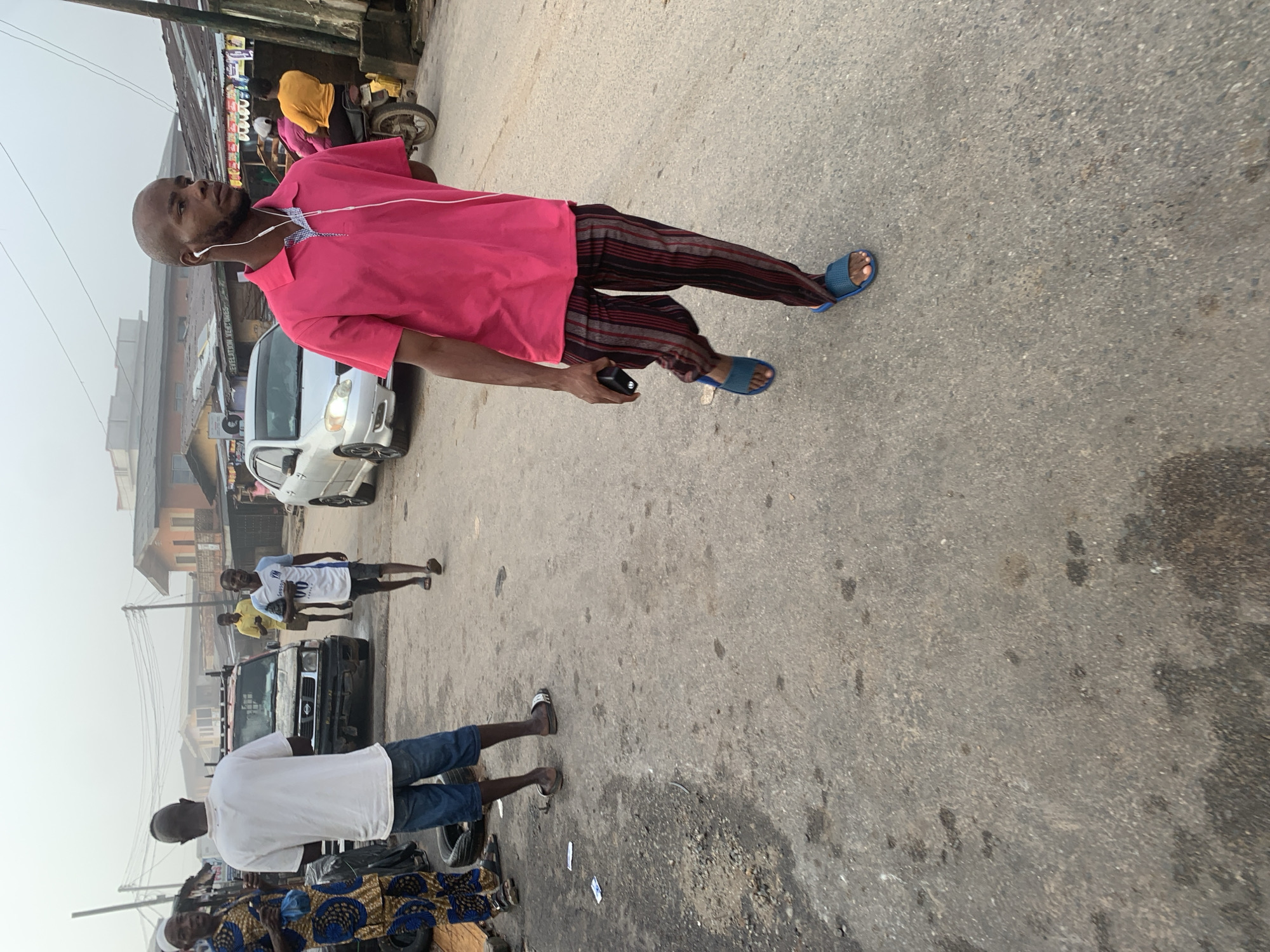 A medium shot captures a street scene in Shagamu, Nigeria, characterized by a dusty, overcast sky and an asphalt road showing signs of wear and tear. In the foreground, a man walks towards the viewer, wearing a bright pink polo shirt and striped pants, with earbuds in his ears and holding a dark object in his right hand. He wears blue slide sandals. Behind him, to his left, another man in a white t-shirt and denim shorts stands facing away from the camera. Further in the background, a white sedan is parked on the right side of the road, and a person on a yellow motorcycle is visible passing by on the right. A black pickup truck is parked on the left side of the road. Several other people, some in traditional clothing, are scattered throughout the scene, suggesting a busy market or residential area. The overall impression is one of a typical day in a Nigerian urban environment.