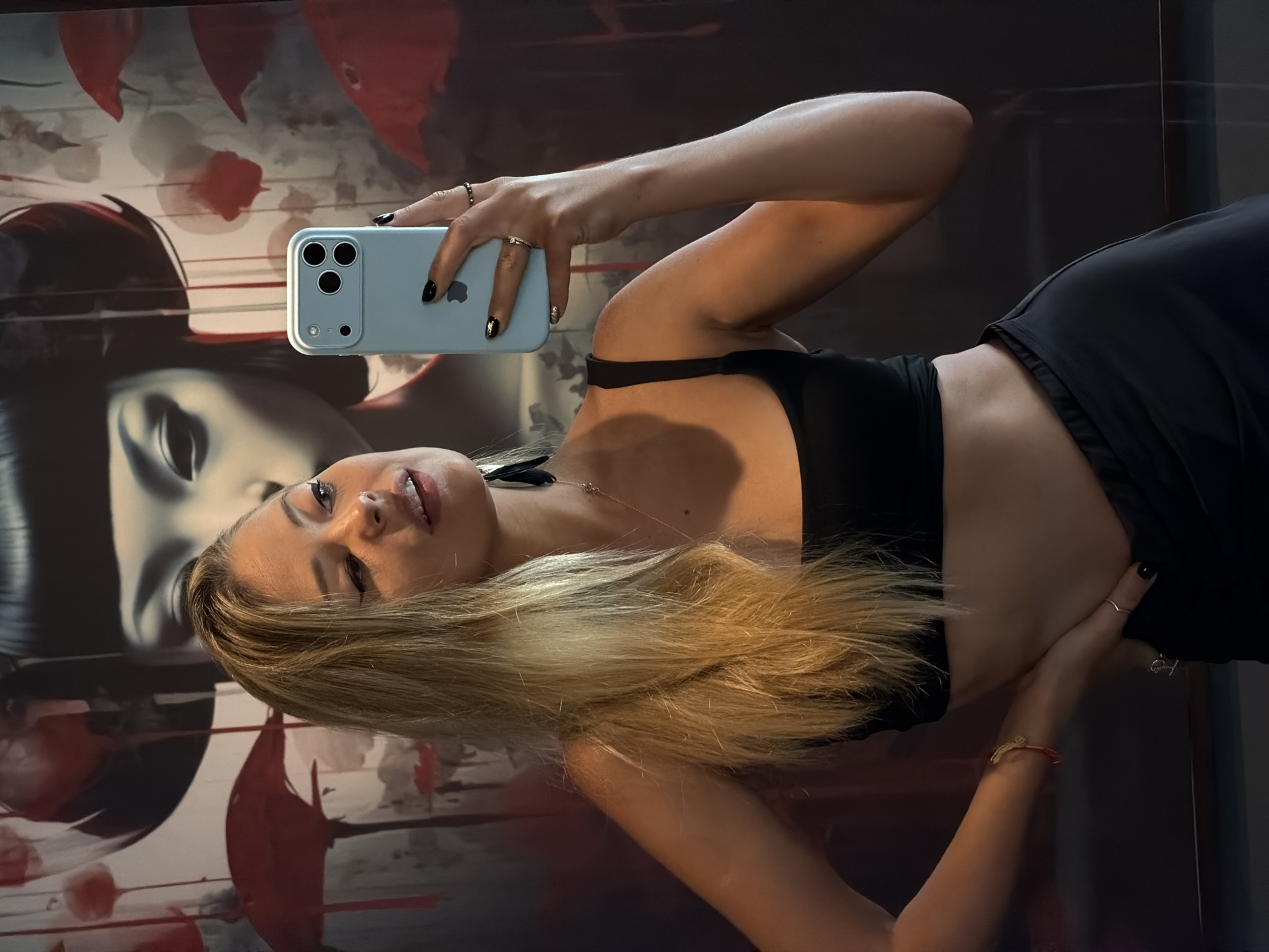 A young woman with blonde hair is taking a selfie in a room. She is wearing a black crop top and black pants. She is holding a light blue smartphone in her right hand. In the background, there is a large painting of a woman's face and red umbrellas. The lighting suggests it is indoors. The exact location within ตำบลเกาะพะงัน, Thailand, is not identifiable from the image alone.