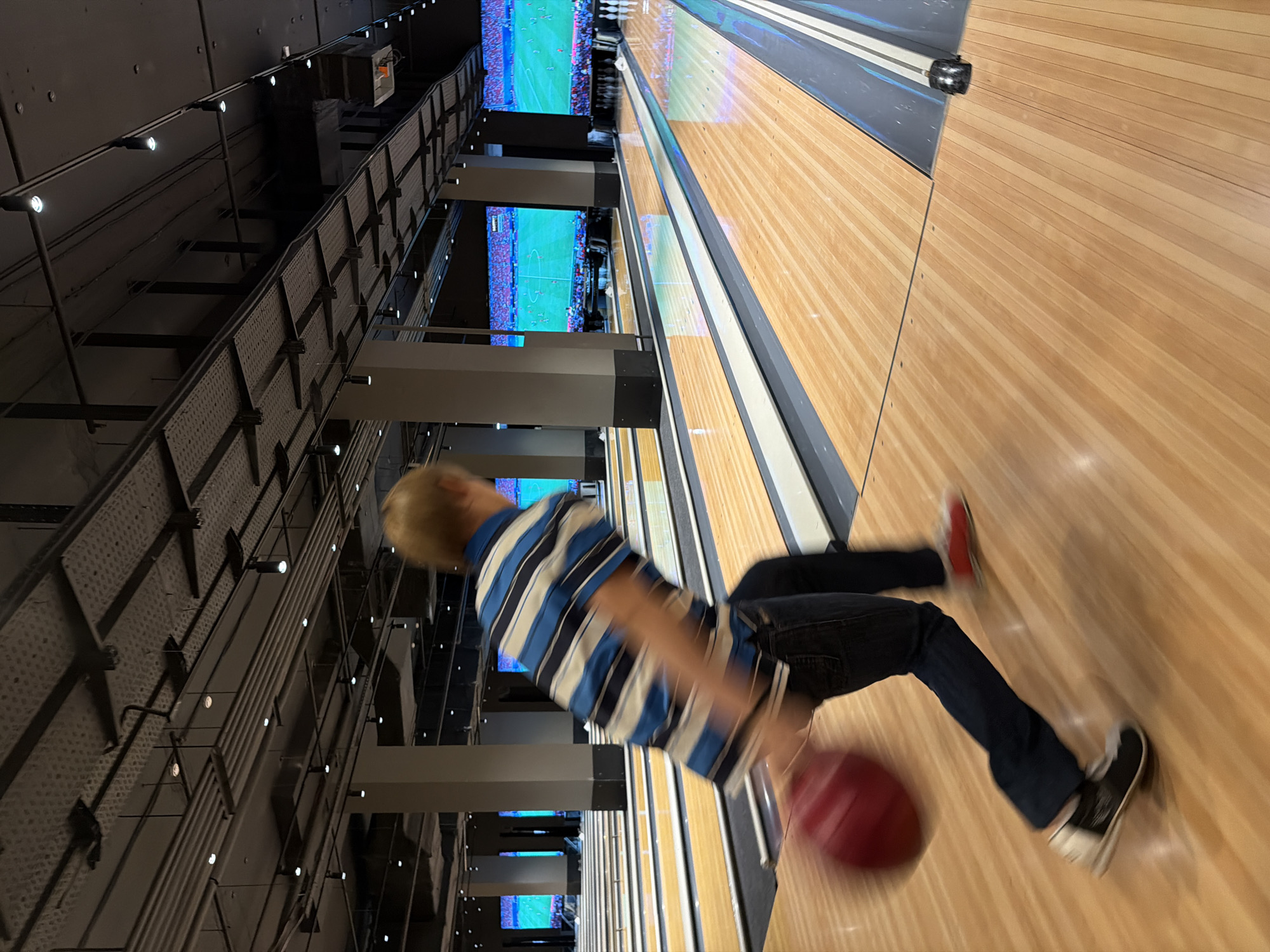 A blurred person is captured mid-action, bowling in a bowling alley. The person, wearing a blue and white striped shirt and dark jeans, is in the process of releasing a red bowling ball. Their movement suggests they are right-handed, with their body leaning forward and their left leg extended. The bowling alley features polished wooden lanes, bordered by dark gray gutters. In the background, large screens display what appears to be a soccer match. The ceiling is industrial, with exposed pipes, conduits, and track lighting. The scene takes place in a modern bowling establishment, potentially in Izhevsk, Russia, indicated by the generic design of a contemporary entertainment venue.