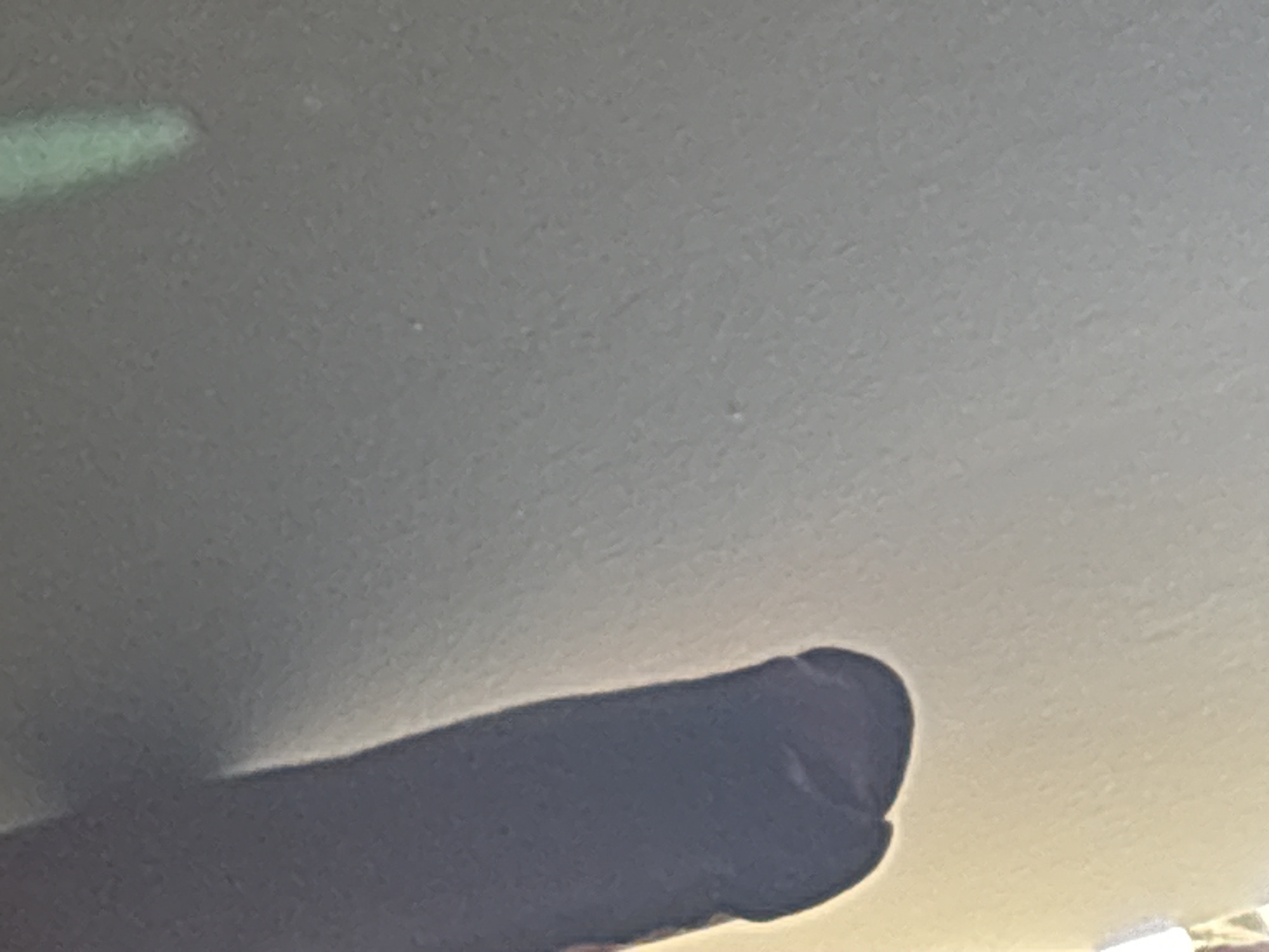 This is a close-up, blurry, and poorly lit image of a dark object on a textured, light-colored surface. The object appears to be a segment of a dark fabric or material, possibly a leg of pants or a sleeve, with some creasing and a rounded bottom edge. There is a faint, blurry green shape in the upper right portion of the image. The overall impression is of an abstract composition with limited detail and unclear context. There is no discernible location in Lugbe, Nigeria, nor any indication of people, actions, or interactions.