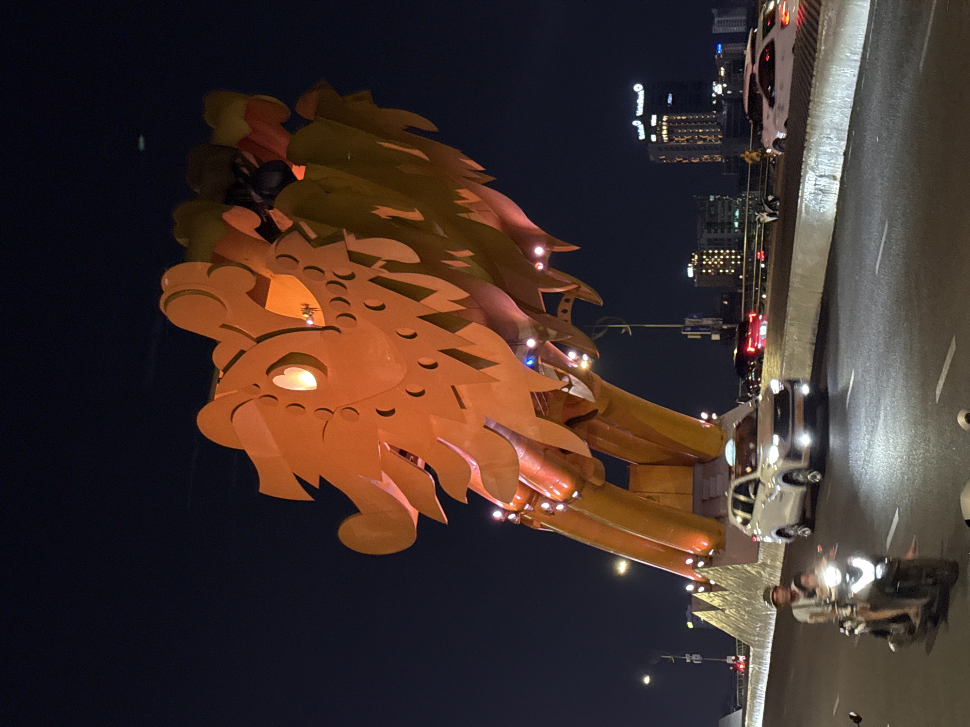 The image captures the iconic Dragon Bridge in Da Nang, Vietnam, at night, with the main subject being the elaborate dragon sculpture. The massive dragon, forming part of the bridge structure, is vividly illuminated in shades of bright orange and yellow, highlighting its intricate scales and fierce, open-mouthed head. Its eyes glow with a warm, bright light, and small blue and white lights are embedded along its body, adding to the decorative illumination.
To the right of the dragon, a multi-lane road, also part of the bridge, is visible with ongoing traffic. Several vehicles, including cars (one prominent white car with bright headlights) and motorcycles, are seen on the road. Reflections of vehicle lights on the dark asphalt suggest a wet or damp surface.
In the background, the nighttime skyline of Da Nang city is visible, featuring numerous illuminated high-rise buildings and hotels. Visible text on one of the background buildings clearly reads "Novotel" and "VINpearl", further confirming the urban setting and location. The scene depicts a vibrant and active urban nightscape. The sky is dark, indicating it is late evening or night.