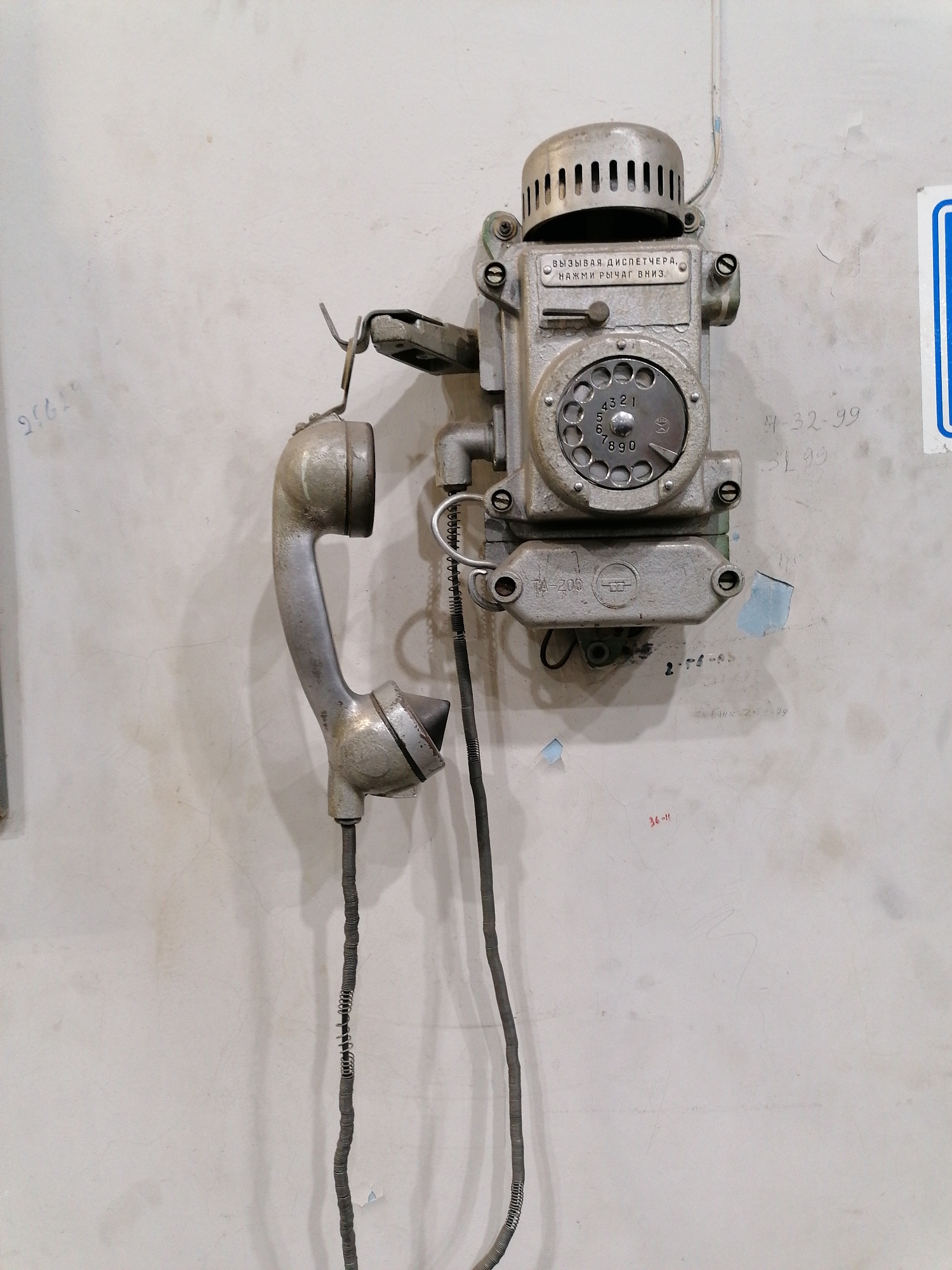 A tarnished, vintage rotary phone is mounted on a weathered, pale wall. The handset hangs loosely, its coiled cord stretched. The phone's metal casing bears the inscription "ВЫЗЫВАЯ ДИСПЕТЧЕРА, НАЖМИ РЫЧАГ ВНИЗ." A rotary dial is centered on the device. Scrawled numbers, possibly phone extensions or dates, are etched onto the wall around the phone. Patches of discoloration and peeling paint mark the wall. The location in the city of Unknown, Unknown cannot be determined from the image.