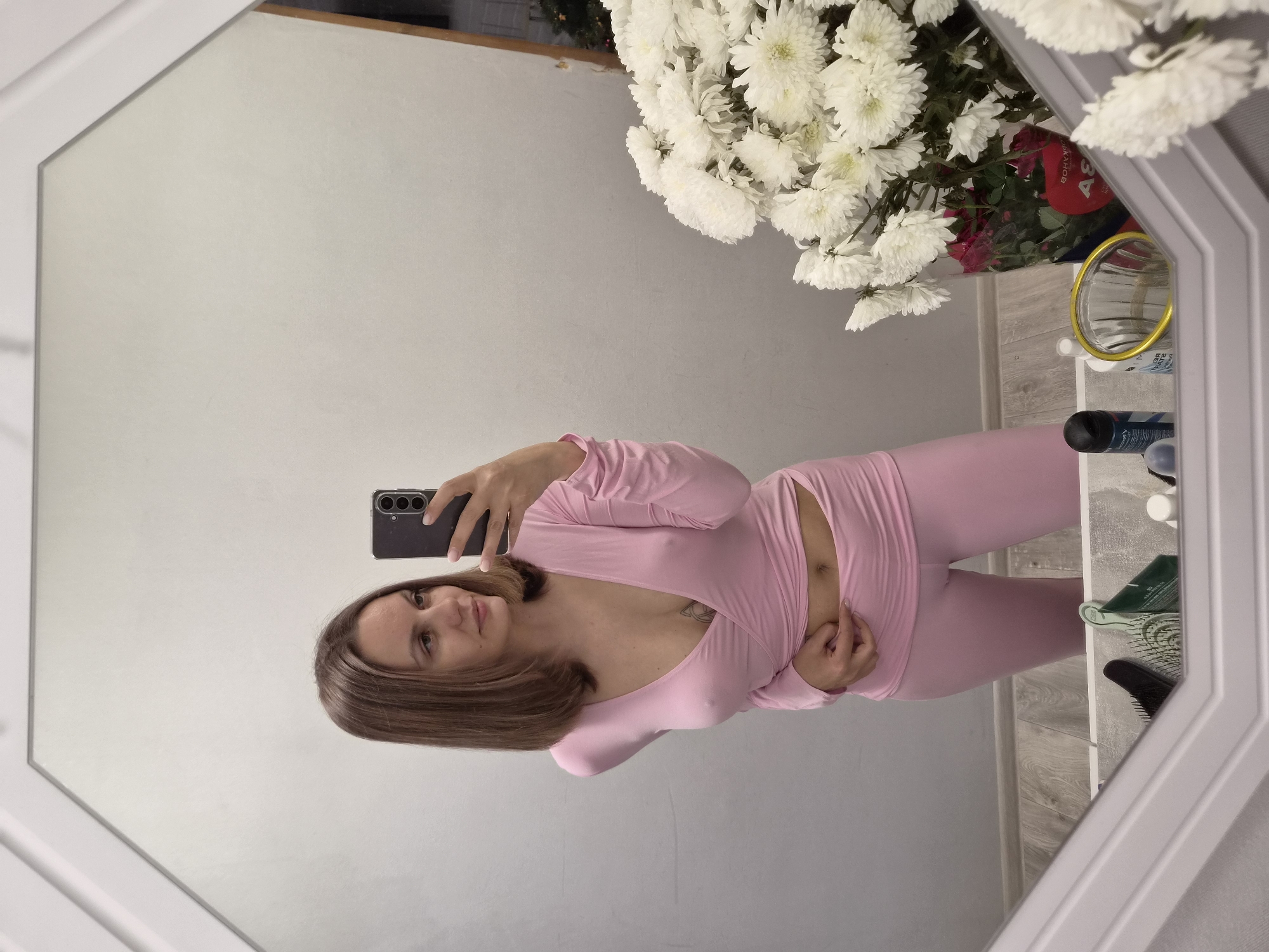 A woman is taking a selfie in a mirror. She is wearing a light pink, long-sleeved outfit. Her hair is shoulder-length and brown. She is holding a black smartphone. The background shows a wall, a bouquet of white flowers with some red roses, and a few personal items on a shelf. The location is Chaikovsky, Russia.