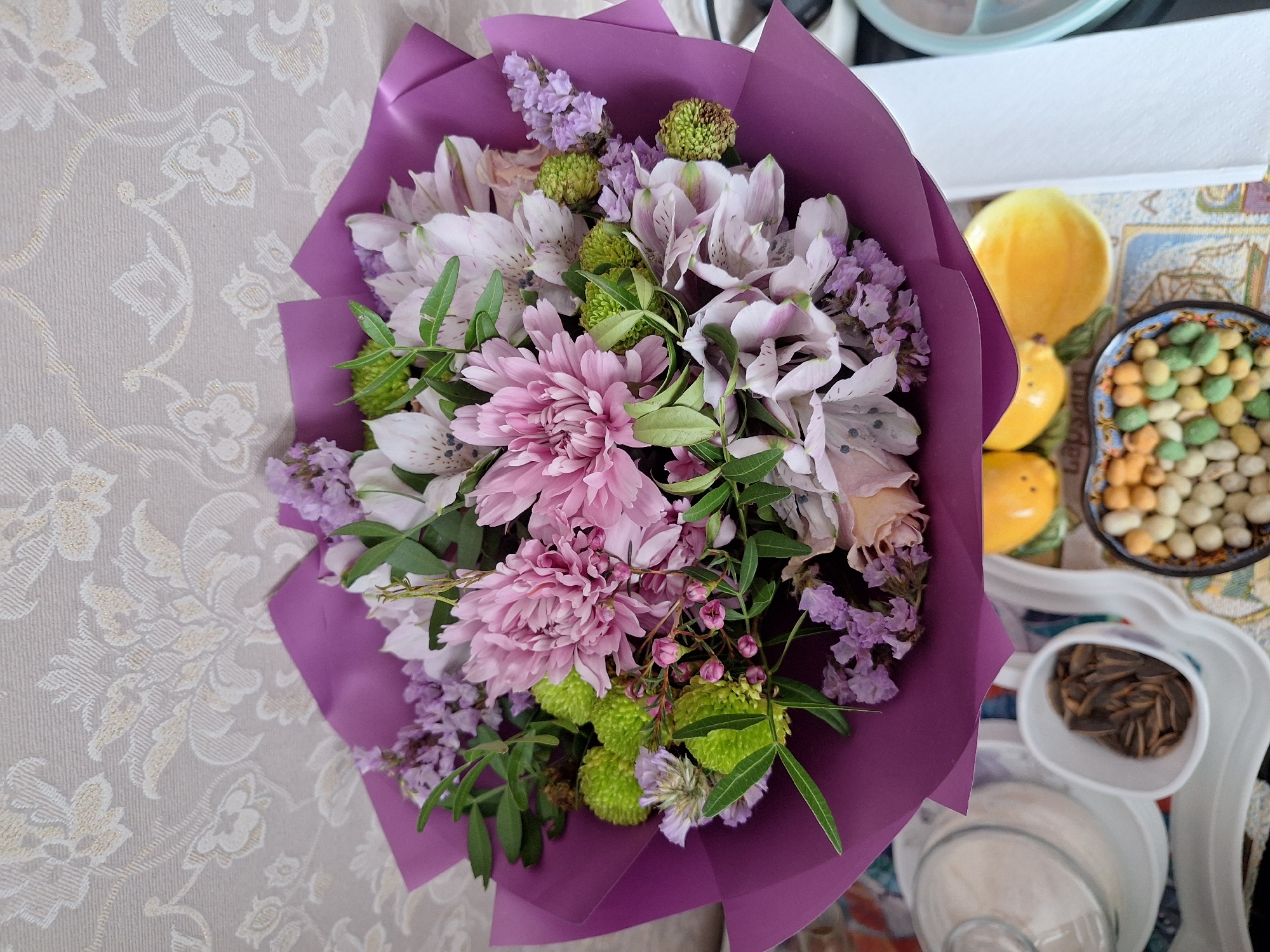 A top-down view of a bouquet of flowers, wrapped in purple paper, sits on a patterned table. The bouquet contains pink chrysanthemums, white and pink alstroemeria, purple statice, and green ornamental flowers. Adjacent to the bouquet, on a white tray, are yellow decorative figurines, a bowl of multi-colored peanuts, a small container of sunflower seeds, and a glass jar with an indistinct white substance inside. The environment appears to be indoors, with a patterned tablecloth and wallpaper visible. No specific location in Moscow, Russia, can be determined from the image alone.