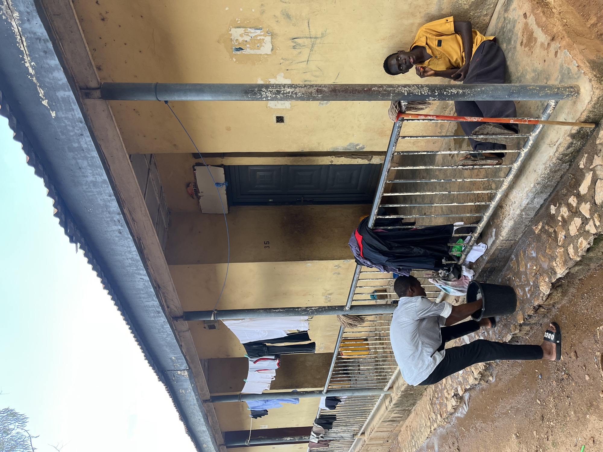 In Jos, Nigeria, an outdoor scene unfolds with a backdrop of a yellow-painted building featuring a corrugated iron roof and a covered walkway supported by metal poles. The walkway is lined with a metal railing, beyond which a person is washing clothes in a black bucket. Various garments are hung on a clothesline strung across the walkway. To the right, another individual is seated on the ground near the railing, looking towards the camera. A mop leans against the railing, and a yellow plastic container is visible behind the railing. The ground is a mixture of dirt and patches of water.